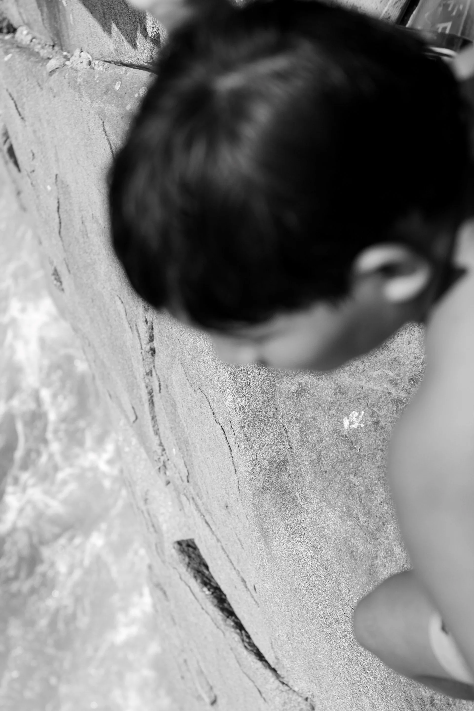 Black and white photo of a person's head with dark hair, seen from behind, focusing on the top of the head and part of the ear, against a concrete wall background.
