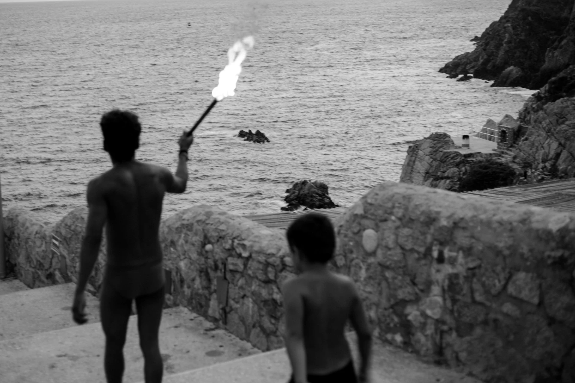 A black and white photo of a coastal scene with three children playing near a stone wall, while a person holds a lit torch near the water in the background.
