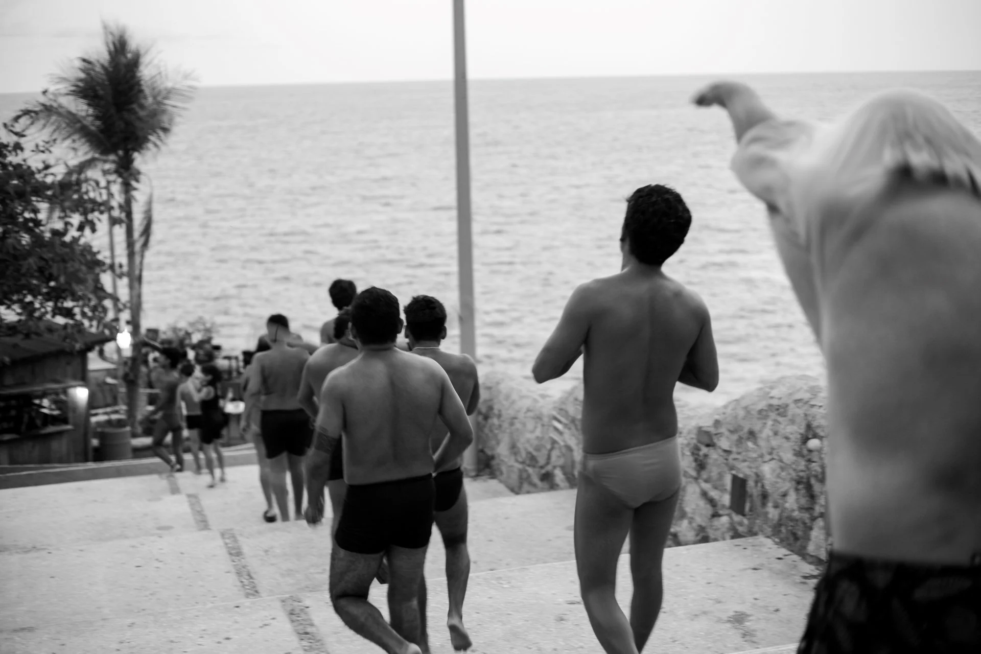 People walking along a beach boardwalk near the ocean, some shirtless, with a DJ playing on a platform, with palm trees and a flag in the background.
