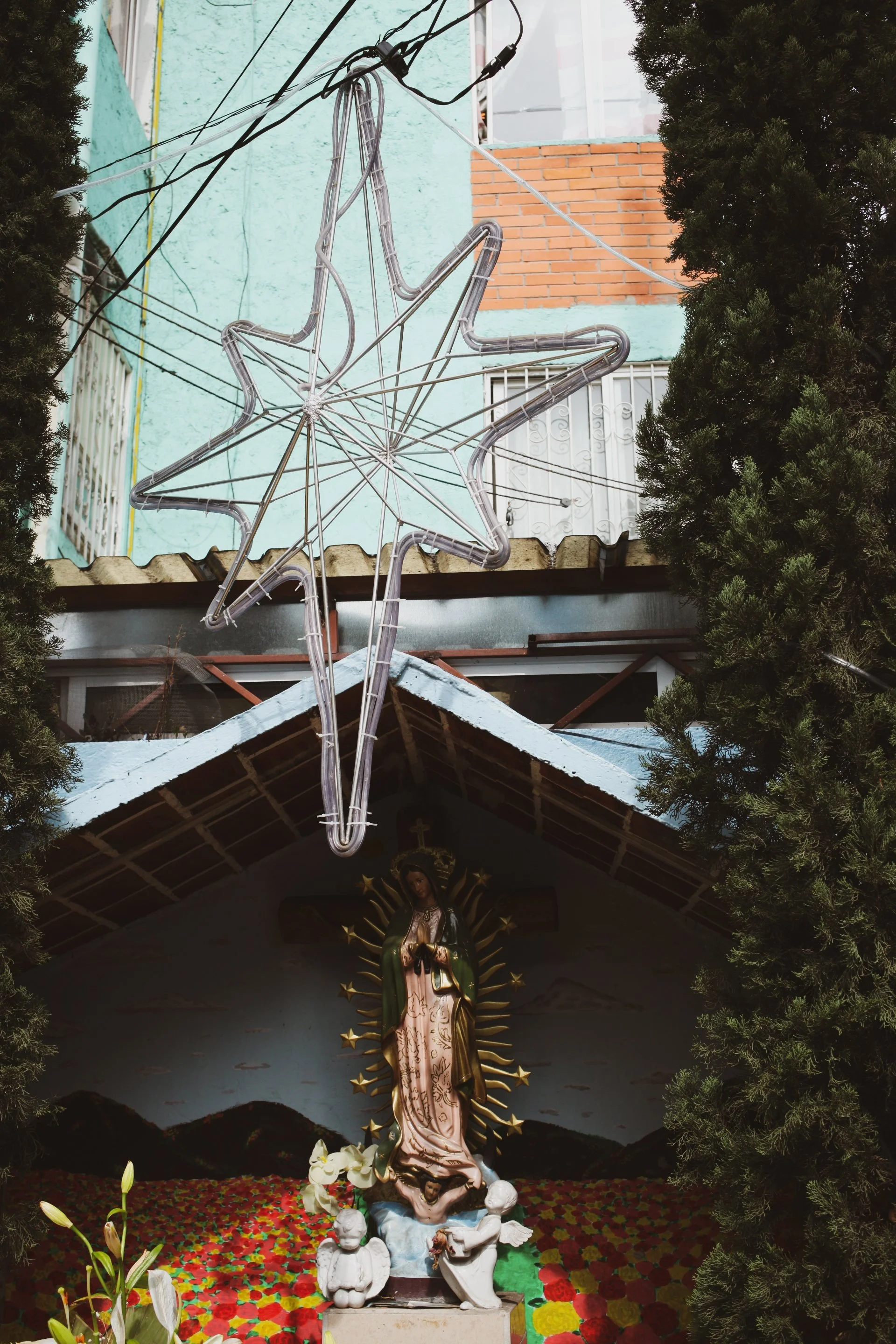 Statue of Virgin Mary surrounded by artificial flowers and greenery, with a star-shaped light fixture hanging above, inside a small church or shrine.