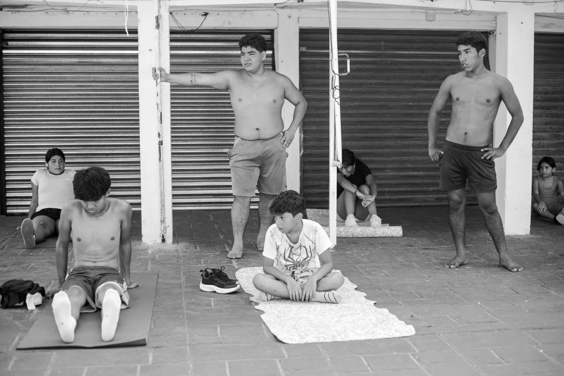 A group of young men and boys are gathered outdoors, some sitting on yoga mats and others standing or leaning against a structure. Several of them are shirtless, suggestive of a fitness or yoga class. The background features closed metal roller shutt
