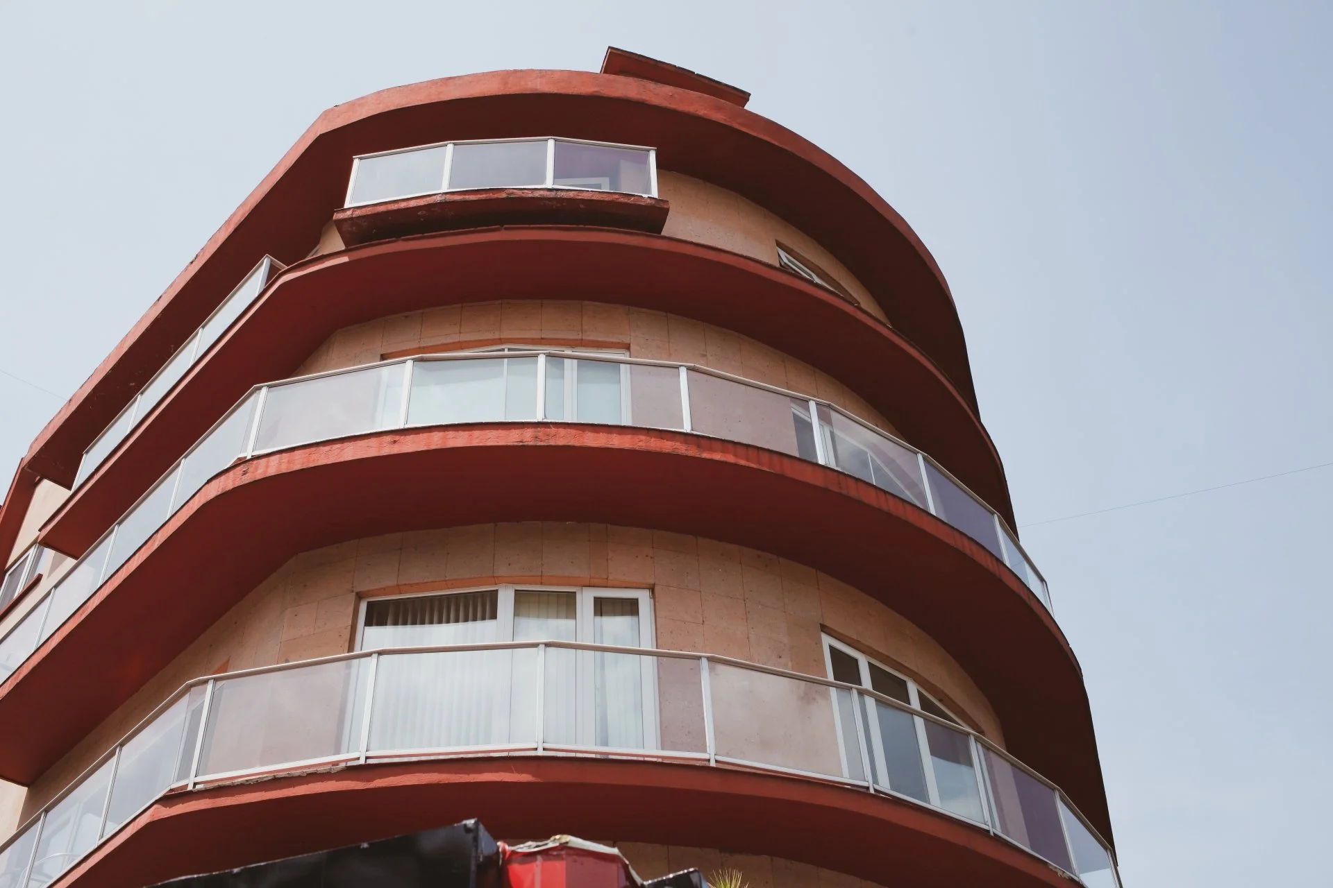 A multi-story building with curved balconies and glass railings, painted in beige and red, against a clear sky.