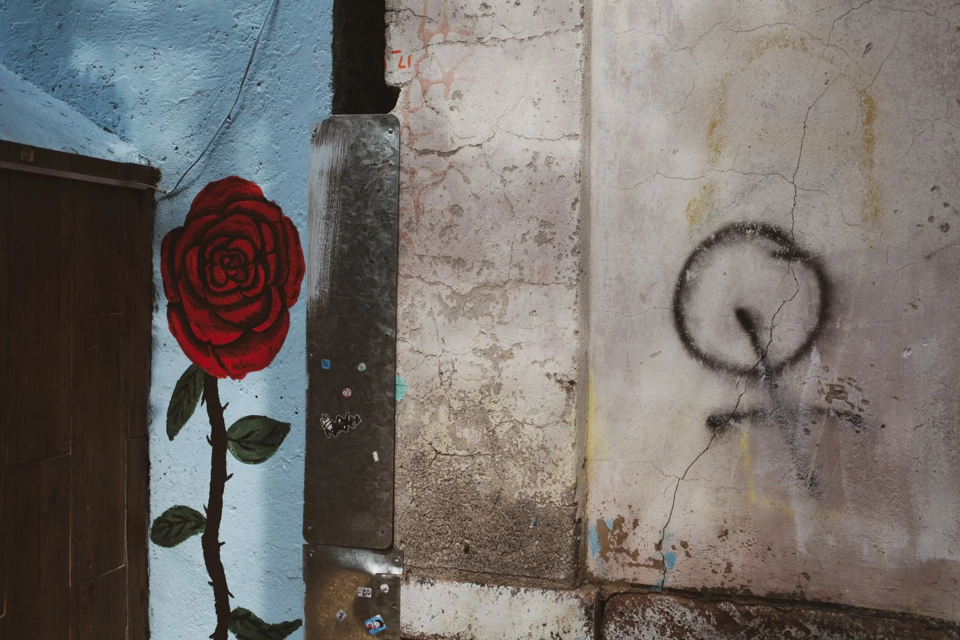 A wall with a painted red rose and green leaves on the left side, a rusty metal bench attached to the wall, and a black spray-painted circle with a line on the right side of the wall, which is cracked and weathered.