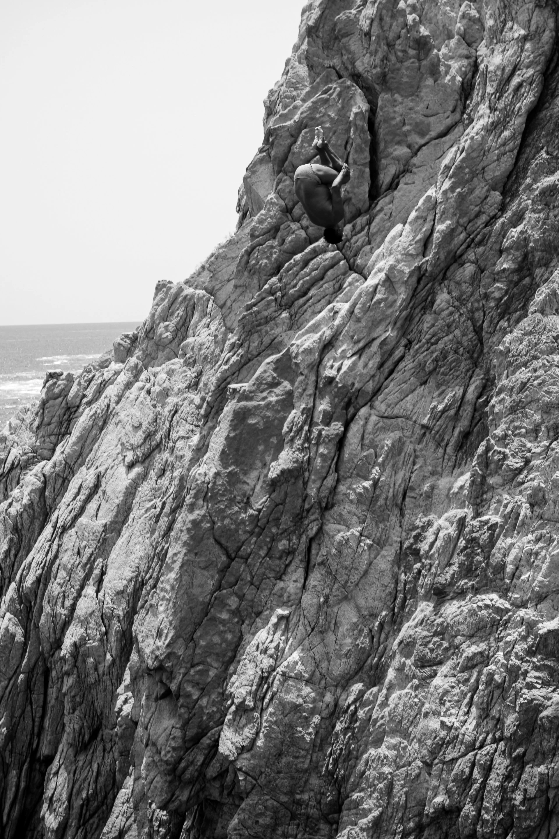 A person in a black shirt and shorts is rock climbing on a rugged cliff face near the ocean.