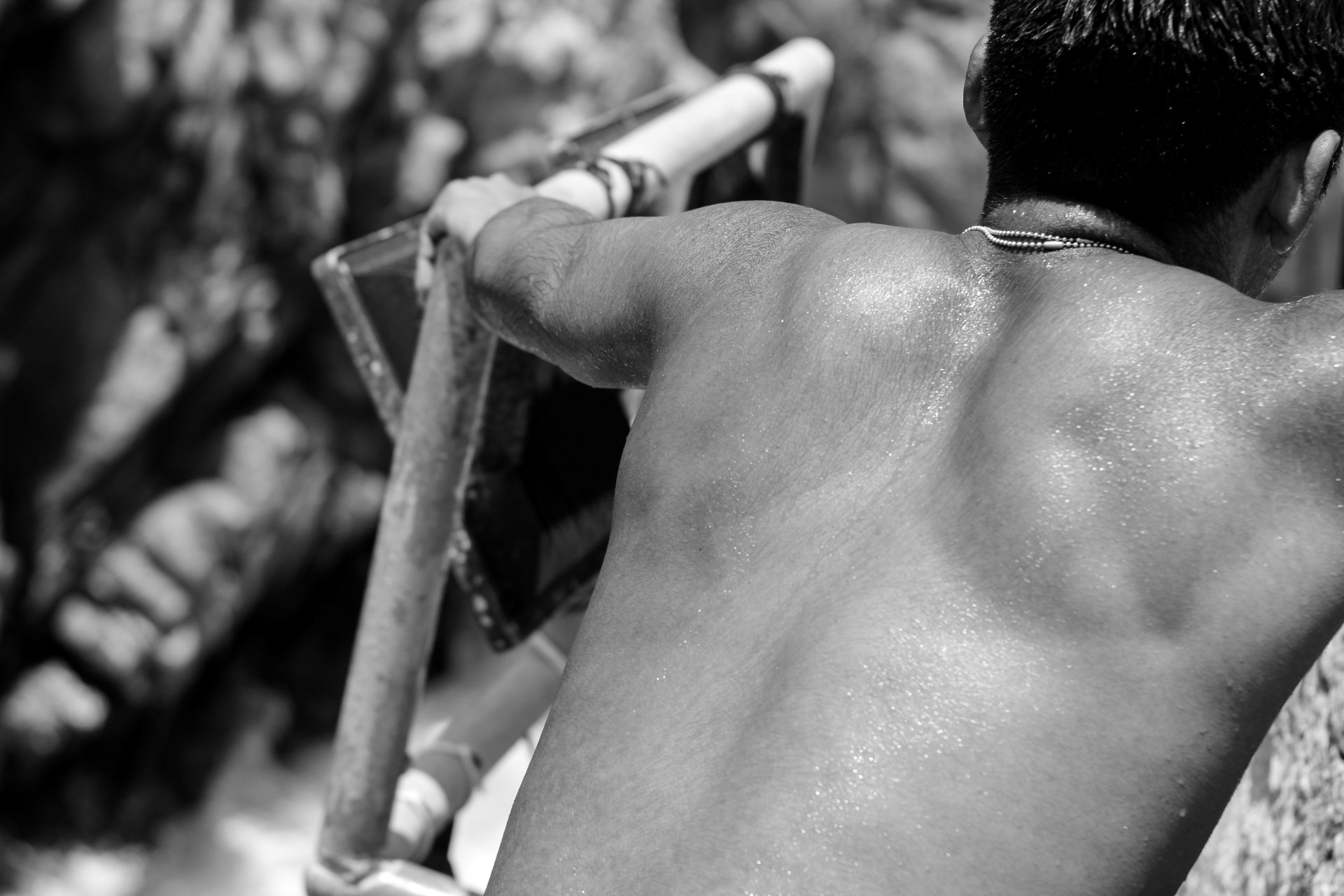 A black and white photo of a person's back, showing lifted arms, part of a strap around the shoulder, and a necklace, with blurred background and natural lighting.