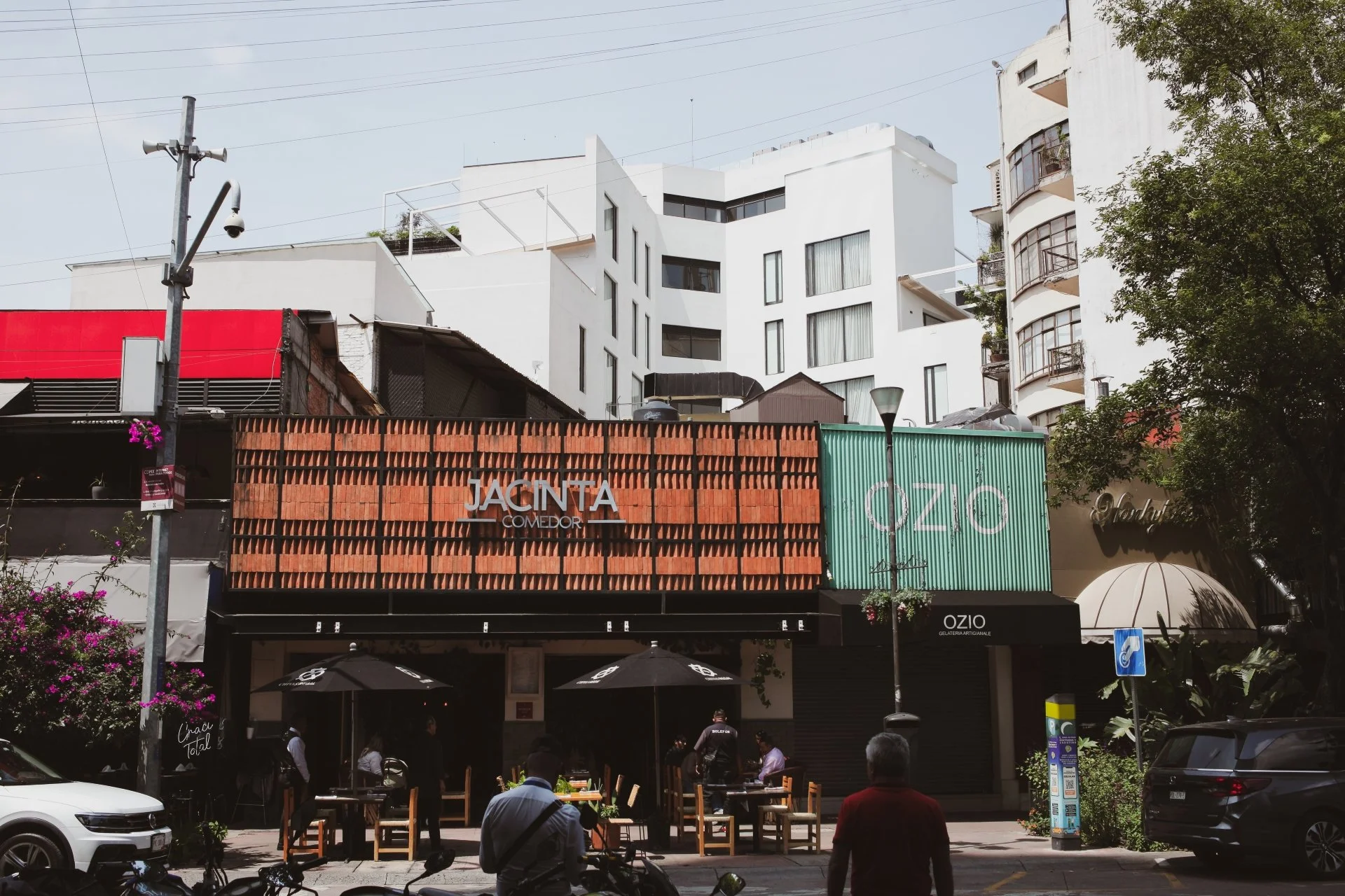 Street view of a building with cafes and restaurants, outdoor seating, parked cars, and people walking. Signage includes 'JACINTA COMEDOR' and 'OZIO'.