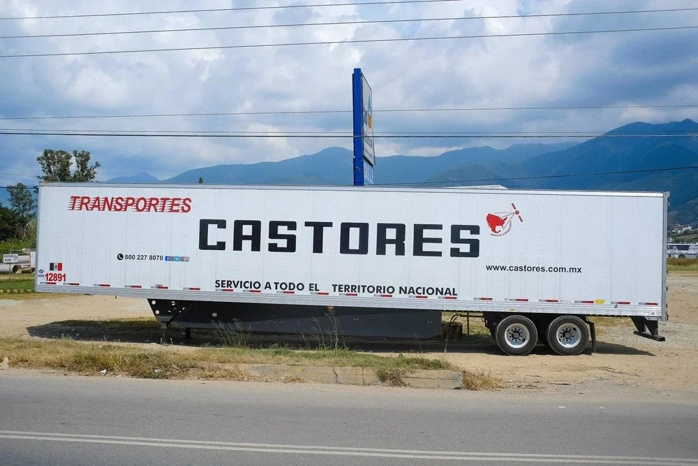 A large white truck trailer with the word 'CASTORES' in bold black letters on the side, parked on a roadside with mountains in the background. The trailer has a red logo of a bird holding a spear and the website www.castores.com.mx.