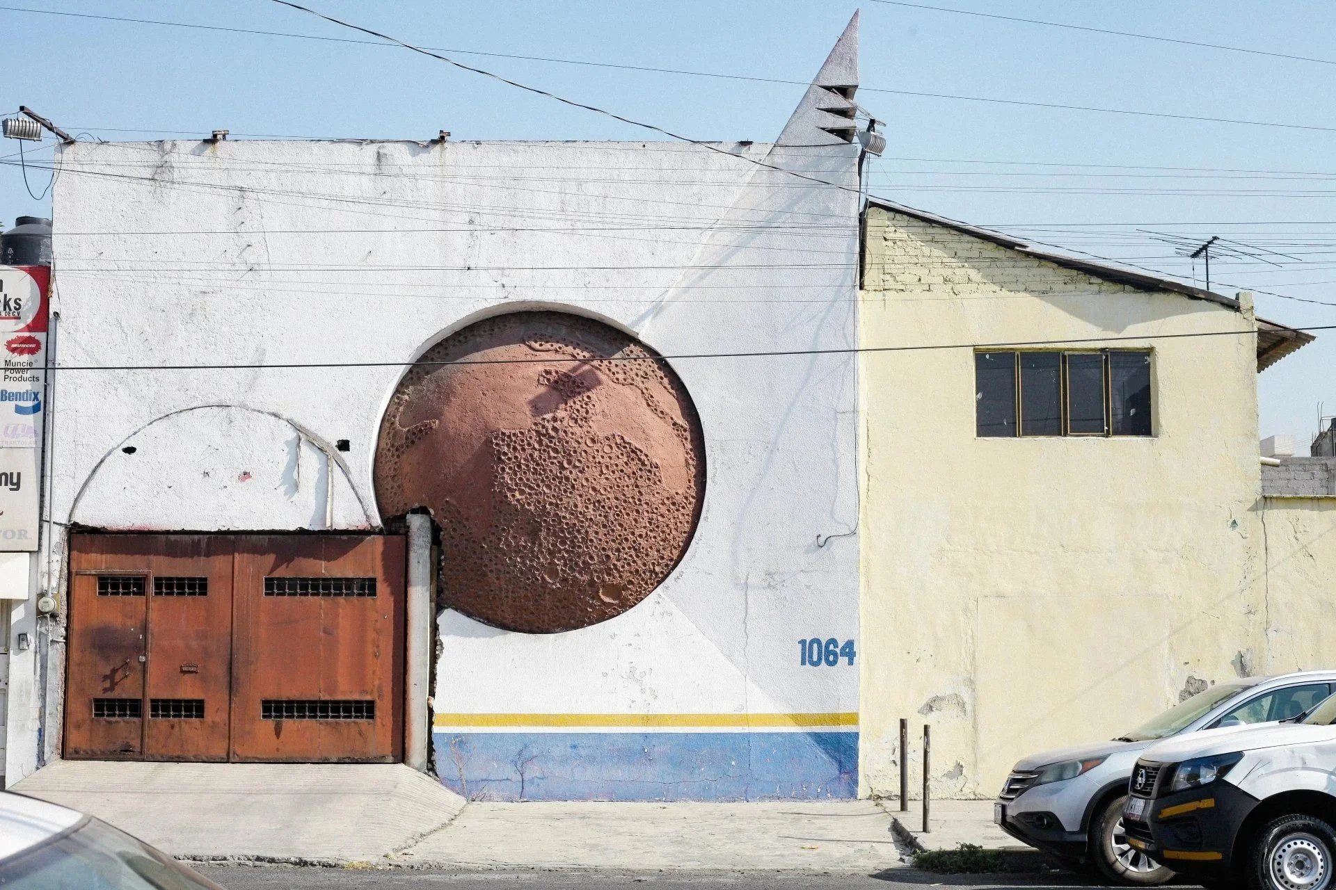 Street view of a building with a large circular art piece resembling a planet with craters, and a wooden garage door. The building has a white and yellow exterior, with a window and roof with antennas. Parked cars are visible in the foreground.