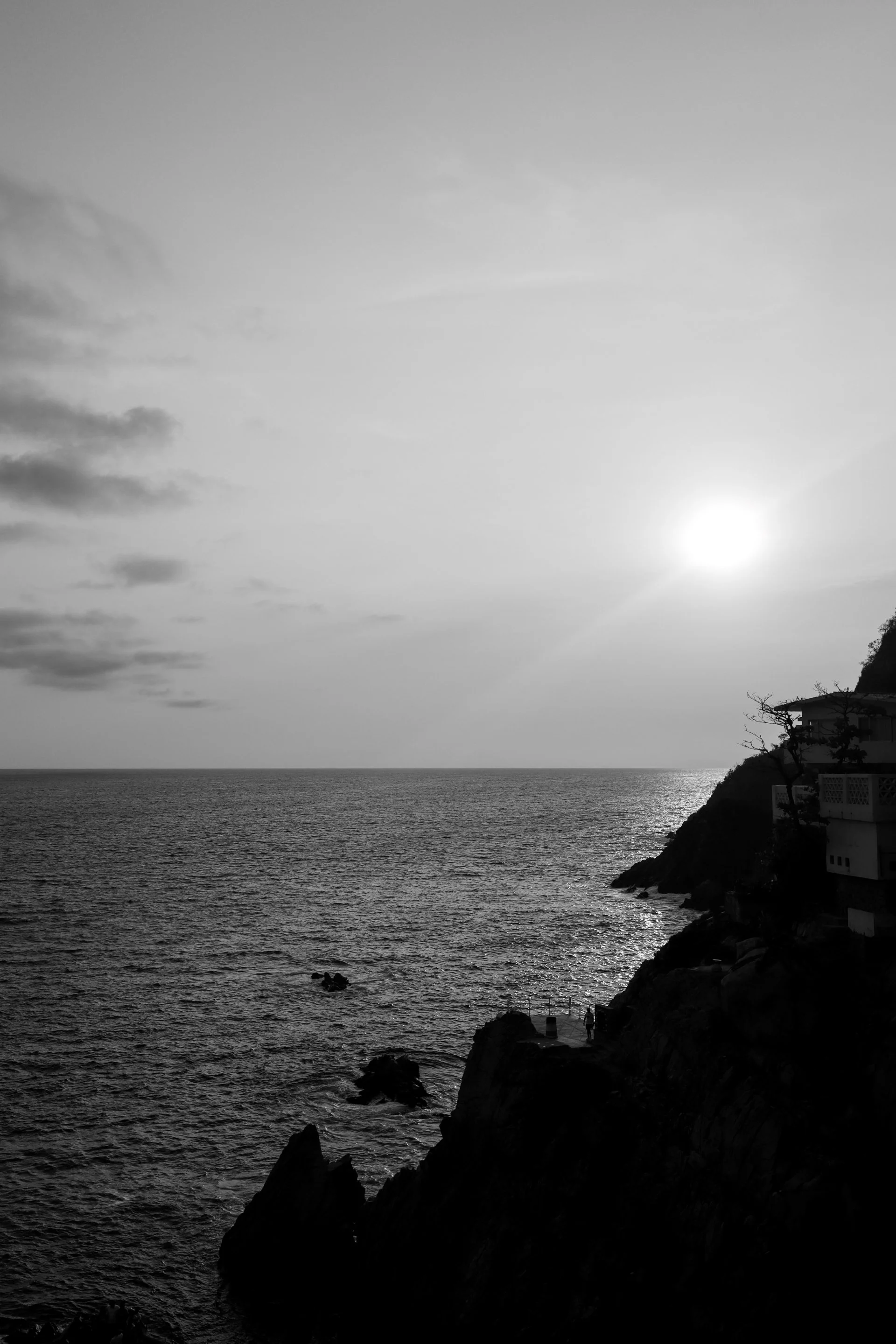 Black and white photo of a sunset over the ocean with a rocky shoreline and silhouetted trees and buildings.