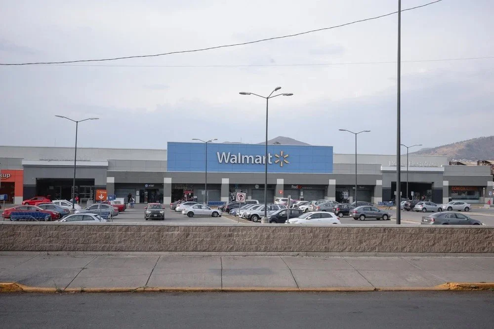 Front view of a Walmart store with a parking lot full of cars, streetlights, and a sidewalk in the foreground, hills visible in the distance.
