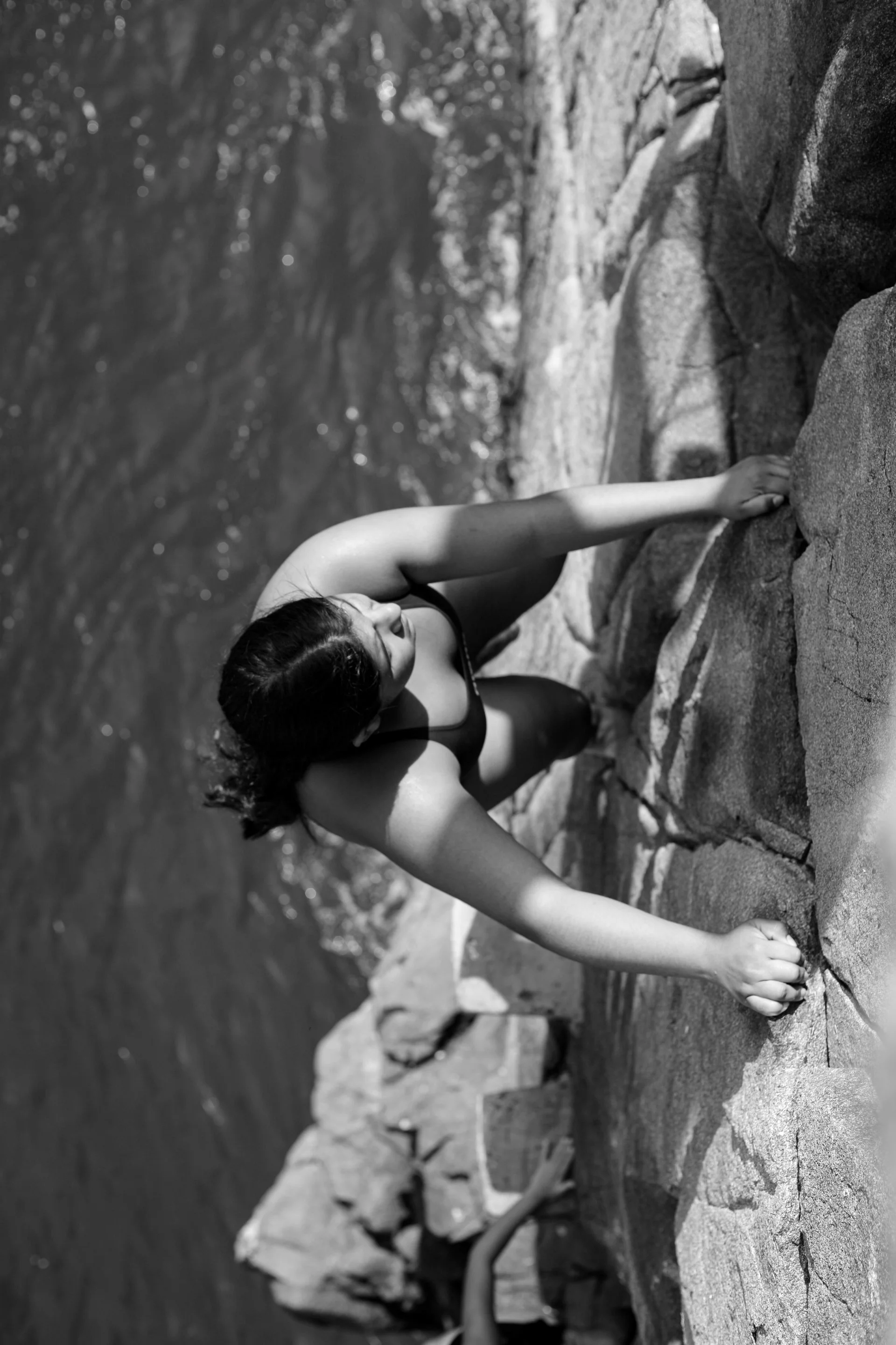 A woman climbing a rocky surface near a body of water with trees in the background, captured in black and white.