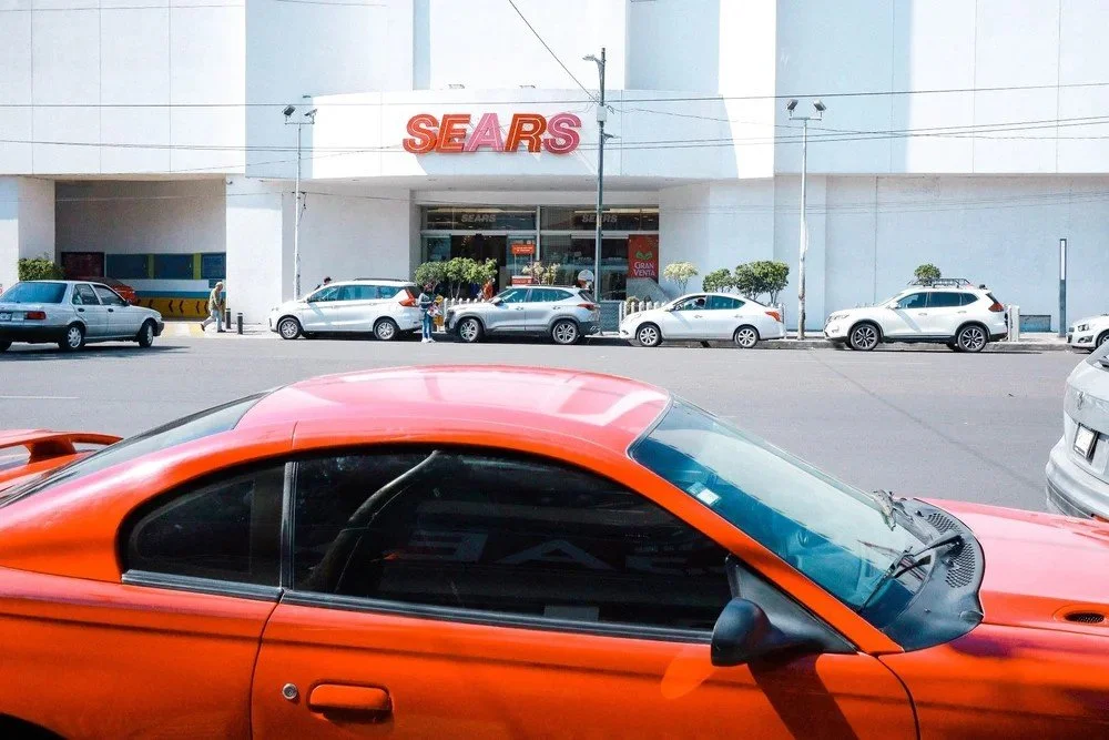 A parking lot outside a Sears store with several parked cars, including an orange sports car in the foreground.