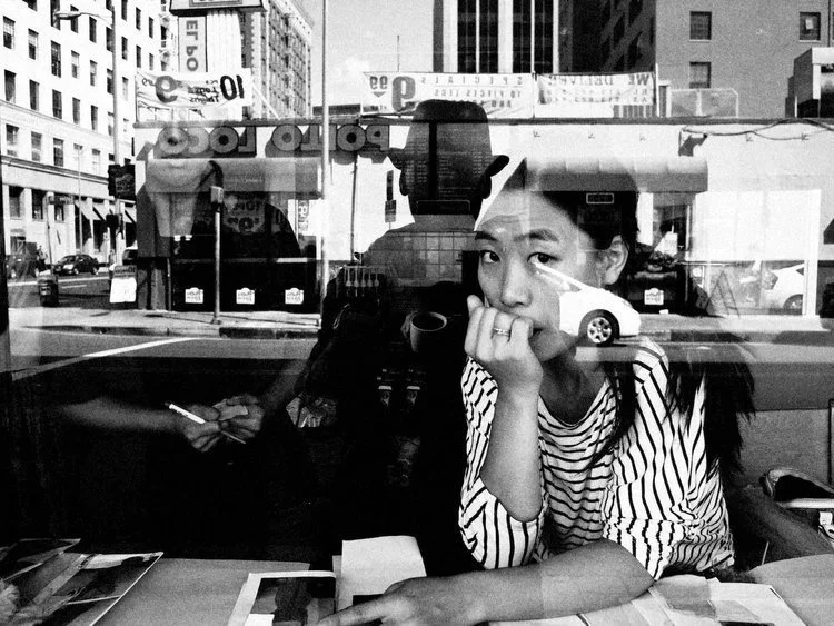 A young woman sitting at a table inside a cafe, looking out of a window with reflections of the city street and buildings on the glass.