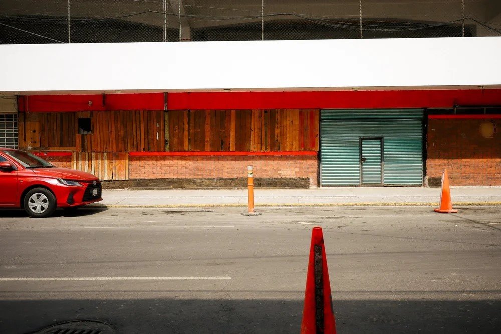 Empty street with a red car parked on the left, construction cones, a closed metal door, and a building with a wooden and brick facade.