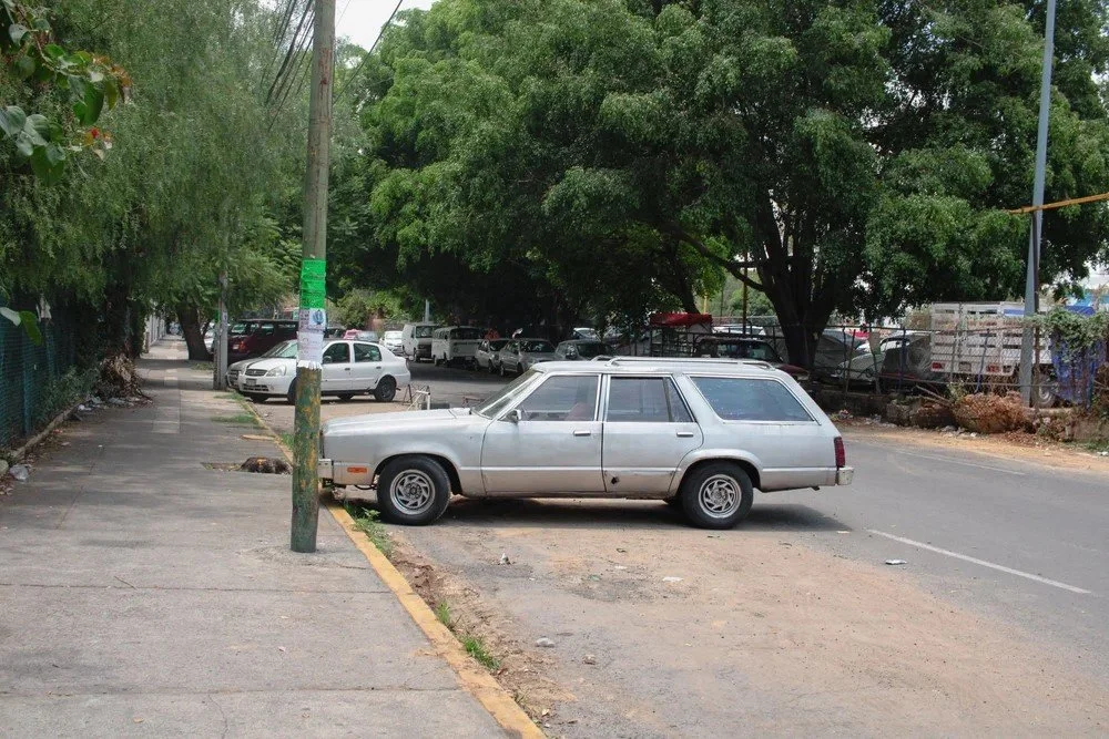 An old silver station wagon parked on the side of the street, partially on the curb and sidewalk, with a tree and other parked cars in the background.
