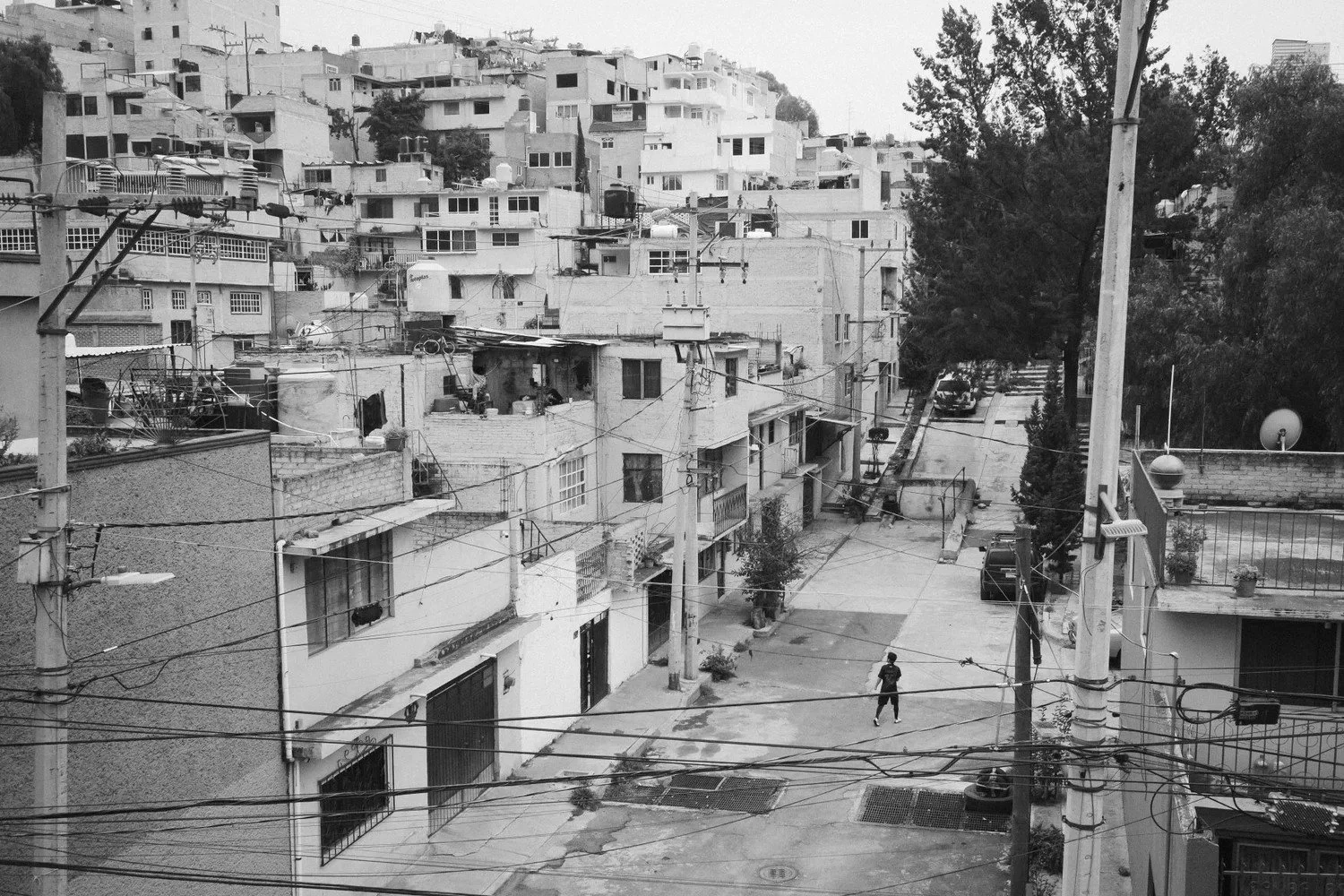 A black and white photo of a densely built urban residential area with multiple multi-story buildings on a hillside. The foreground shows a street with power lines overhead, a person walking, parked cars, and trees lining the street.
