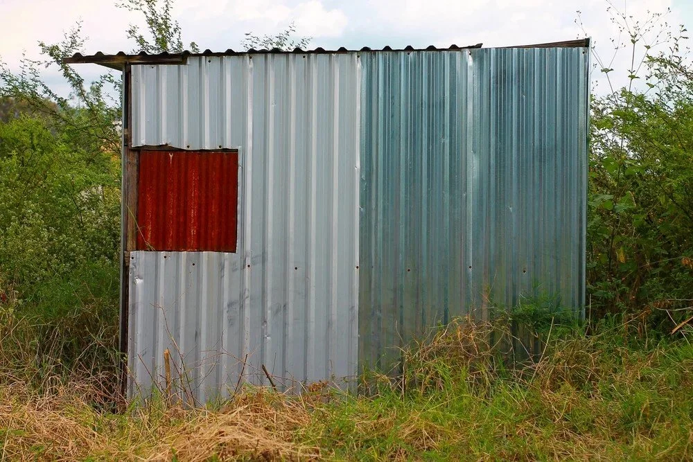 A weathered metal shed with a rusty red window, surrounded by overgrown grass and shrubbery.