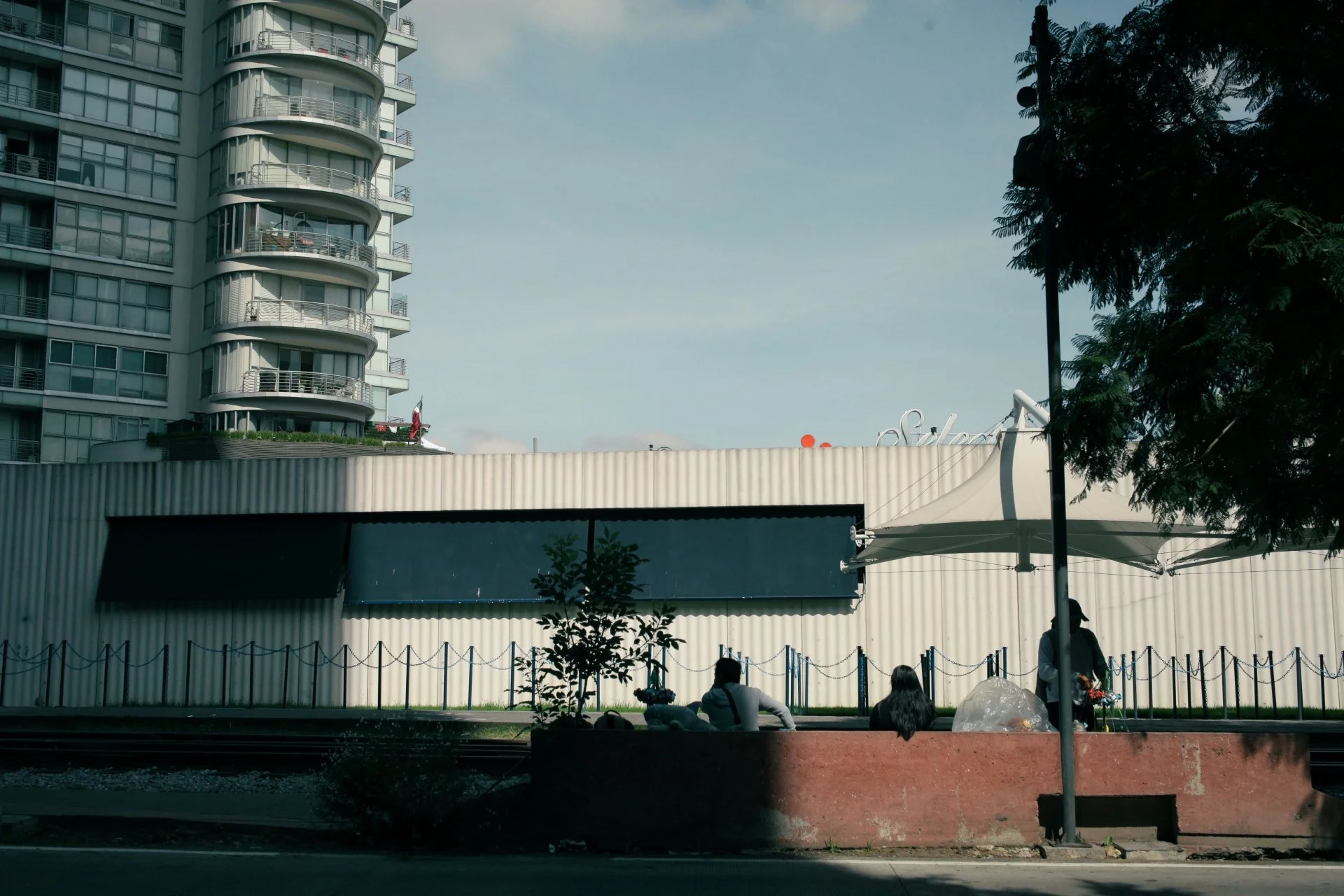 A city scene with a tall modern apartment building on the left, a commercial building with a white canopy on the right, and some people sitting under a tent-like shade in the foreground.