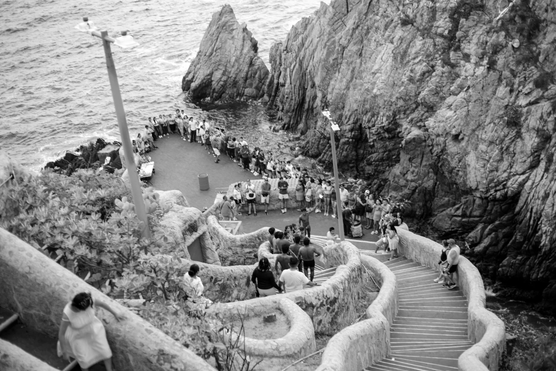 People walking down curving staircase along rocky cliffside near the ocean in black and white.