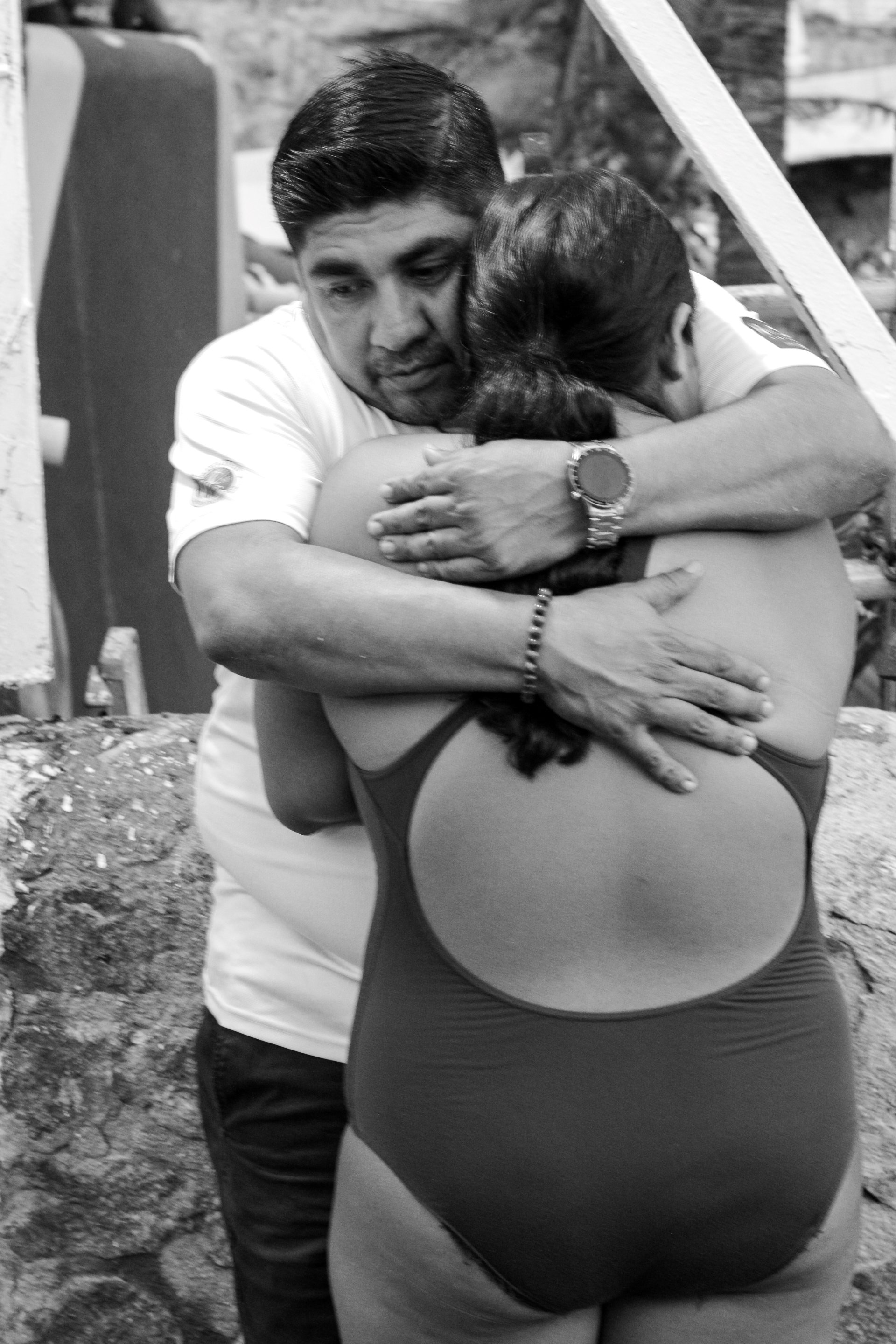 A black and white photo of a man and woman embracing each other outdoors. The man is holding the woman warmly, and both are wearing casual clothing. The background shows some outdoor elements like rocks and a wall.