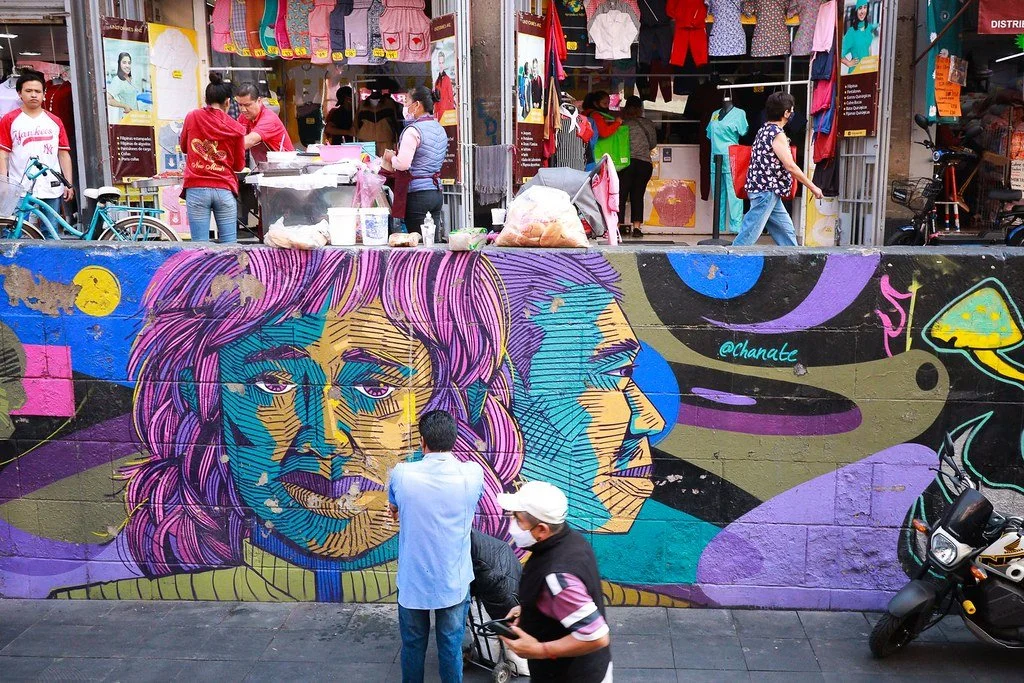 Colorful graffiti mural of two faces on a wall with a busy storefront and pedestrians in the background.