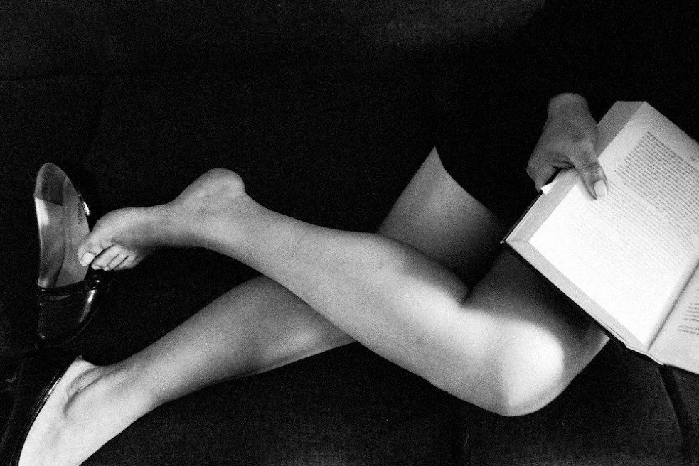 Person sitting on a dark surface, holding an open book and resting their legs over a pair of glasses.