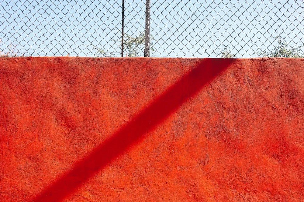 A red textured wall with a diagonal shadow, topped by a chain-link fence and a blue sky in the background.