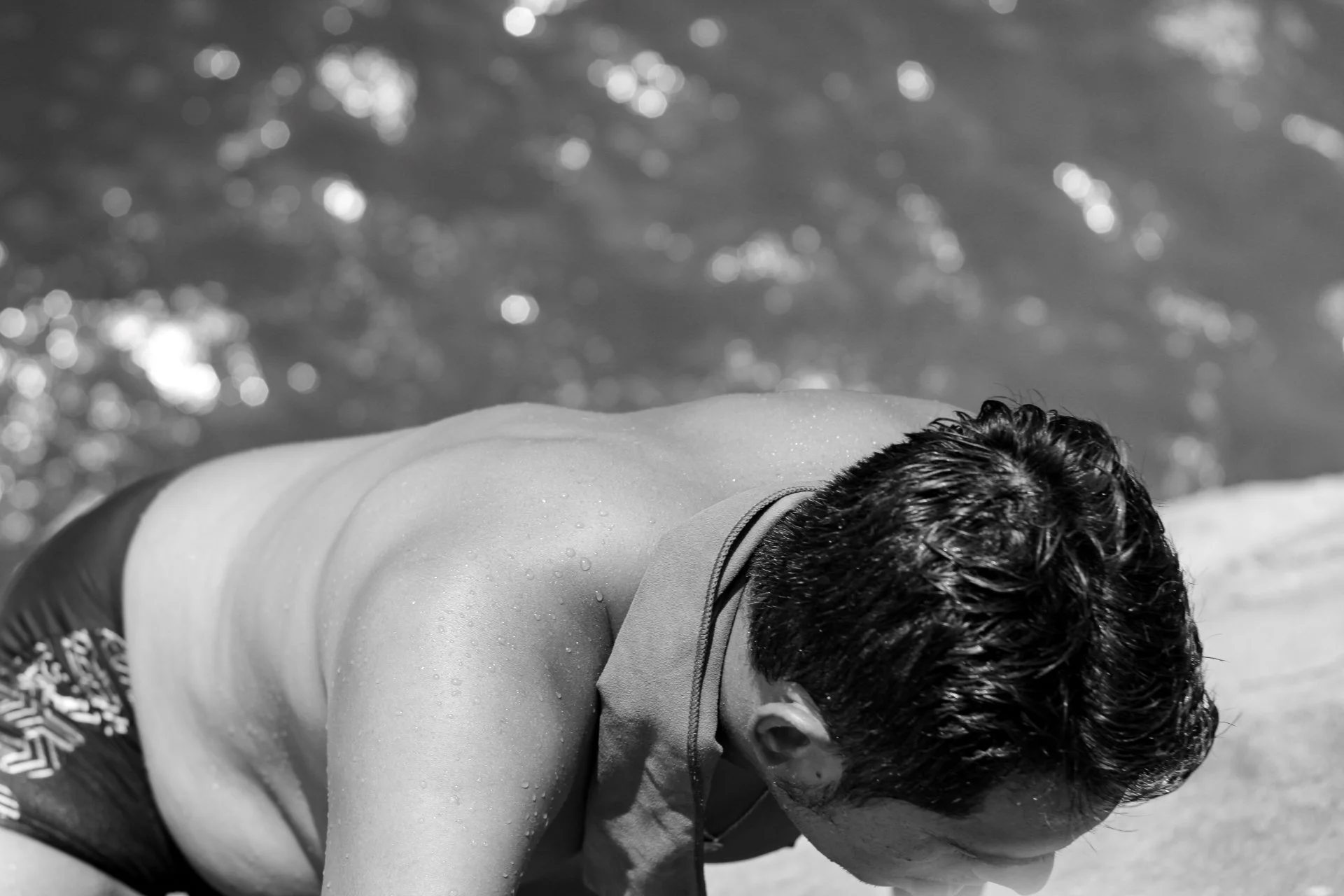A person with dark, wet hair lying face down on a surface, with water droplets on their back, near a body of water with shimmering reflections.