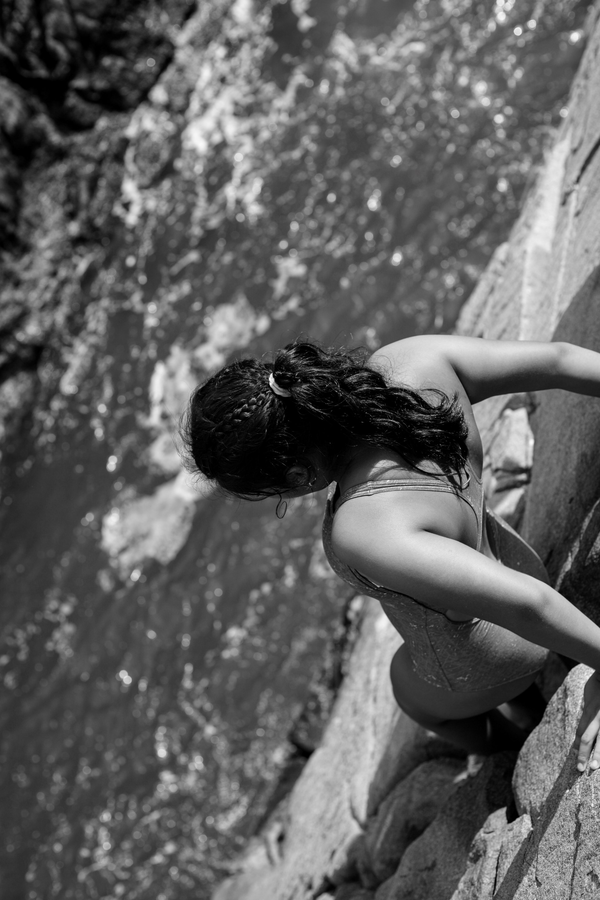 A person with braided hair, wearing a swimsuit, sitting on rocks beside a body of water, with trees and rocks in the background.