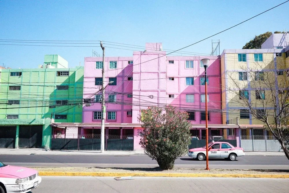 Colorful apartment buildings painted in pastel green, pink, and yellow along a city street with a pink taxi and a pink car in the foreground.