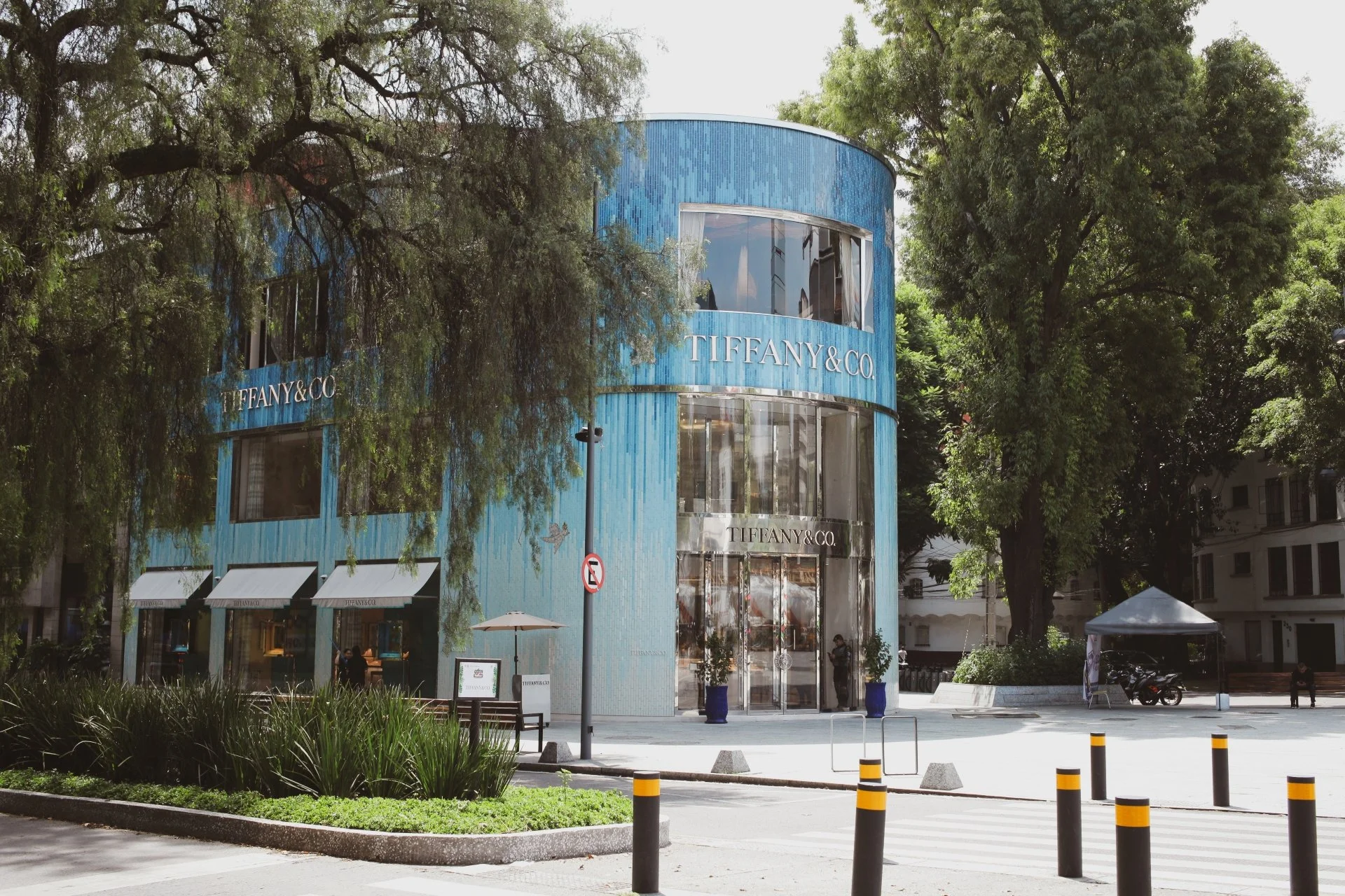 The Tiffany & Co. building in a city setting with lush trees, a sidewalk, a small outdoor seating area with umbrellas, and a parked motorcycle. The building has a rounded facade covered in blue tiles and large glass windows.