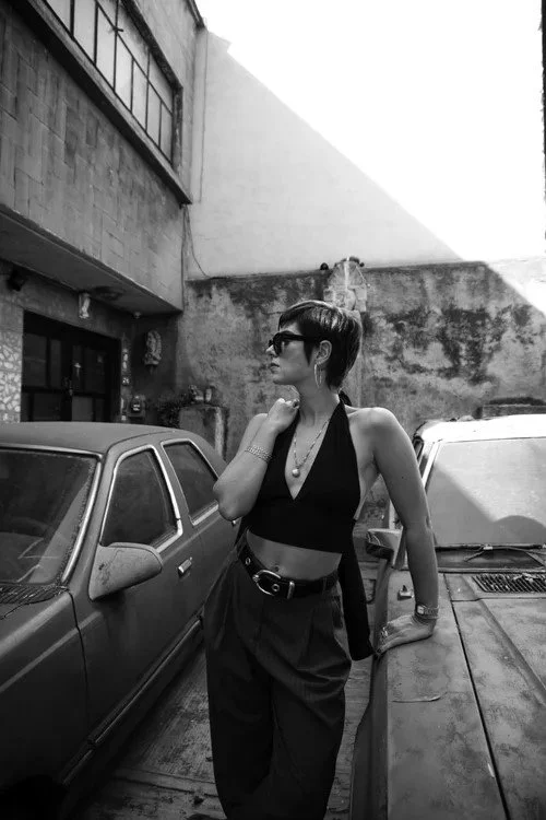 A woman with short hair and sunglasses leaning against a car in an urban alley, wearing a black crop top, high-waisted pants, and jewelry, with a gritty concrete wall and building in the background.
