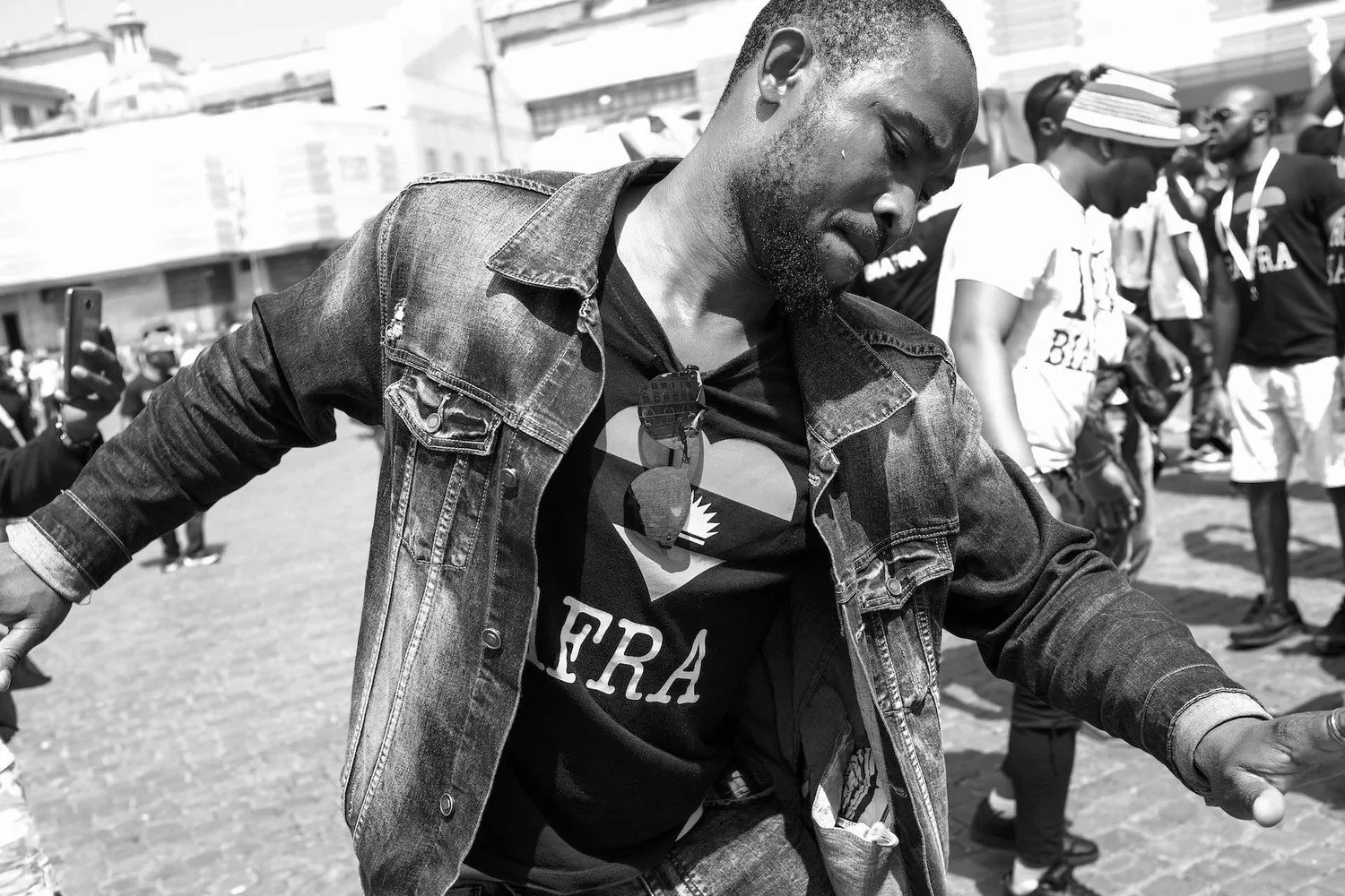 A man in a denim jacket dancing or moving at an outdoor event, with other people in the background.