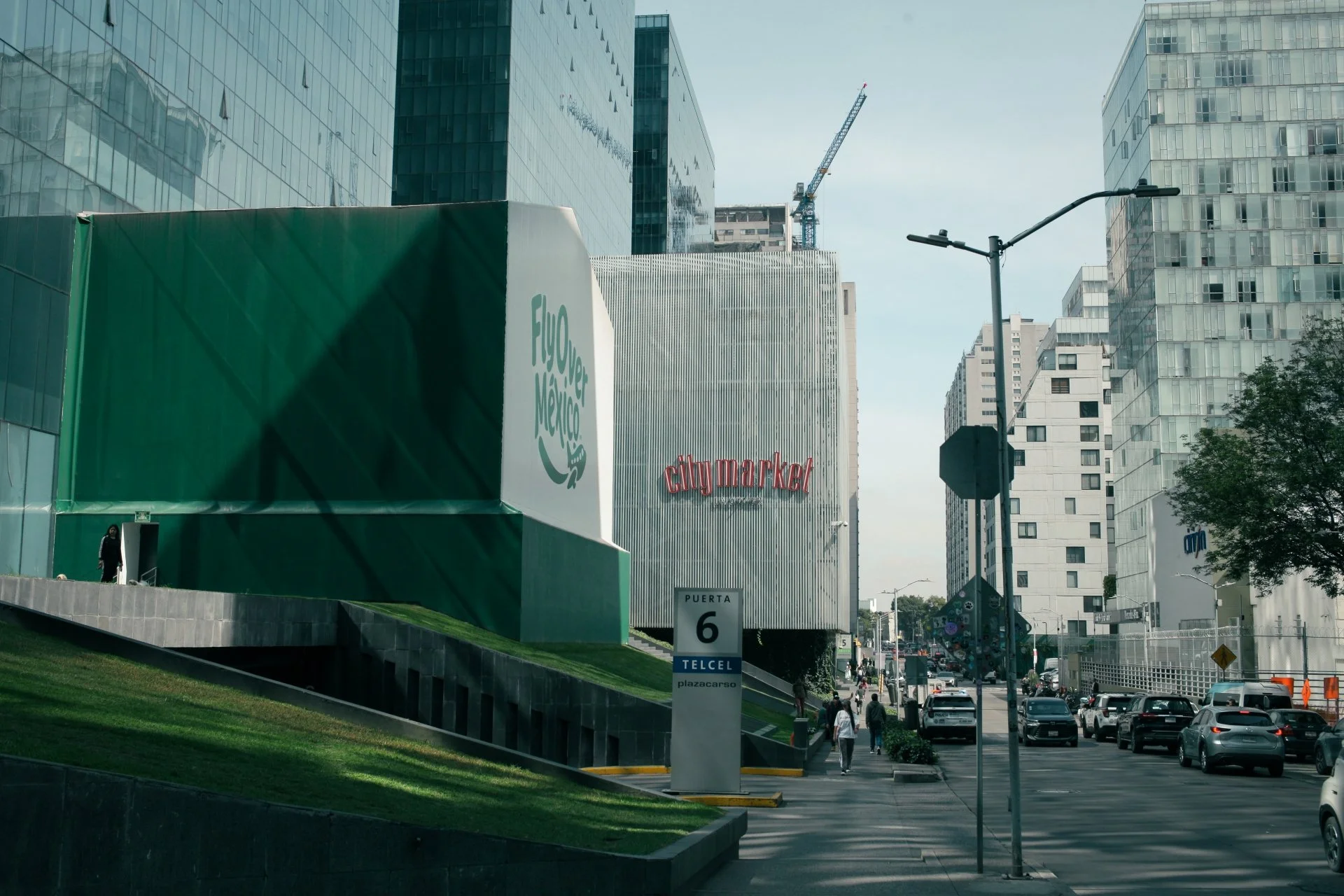 City street scene with modern glass buildings, a large green structure with the logo 'Flyer Mexico,' a sign that reads 'city market,' and multiple pedestrians and cars on the road.