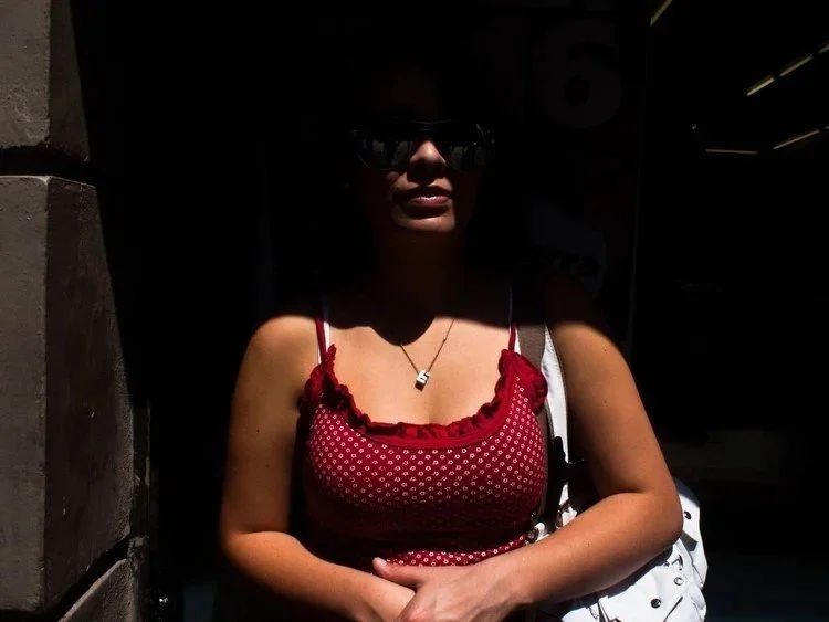 A woman with dark hair wearing sunglasses, a red polka dot dress with ruffle details, and a necklace, standing outdoors in bright sunlight with shadows on her face.