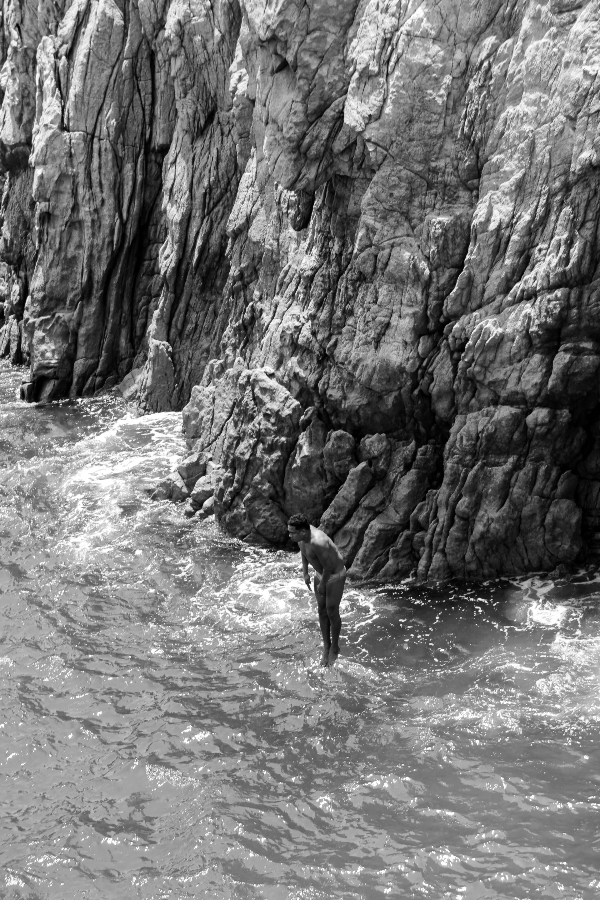 A person standing in the water near a rocky shoreline with large rocks and cliffs behind them.