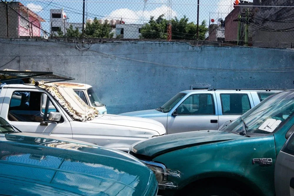 Several parked cars including a green pickup truck, a white SUV with a cloth draped over its windshield, a silver sedan, and a blue vehicle, in an outdoor lot with a gray wall and barbed wire fence, under a partly cloudy sky.