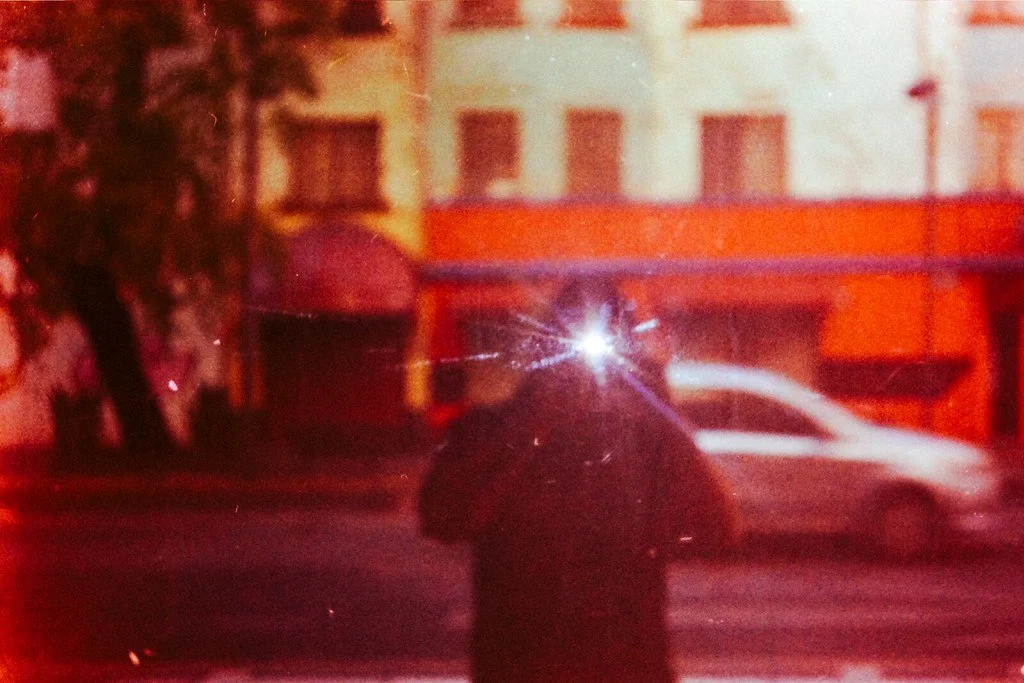 A person taking a photo in a city street at night, with a bright flash reflecting off the camera. Buildings and a passing car are visible in the background.