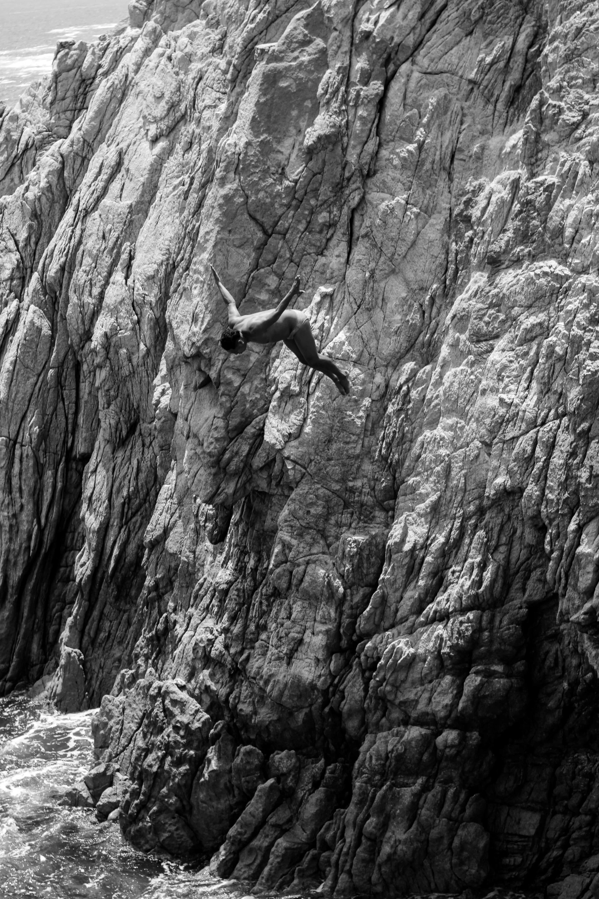 A person rock climbing on a rugged rocky cliff near the ocean, with waves visible at the bottom of the cliff.