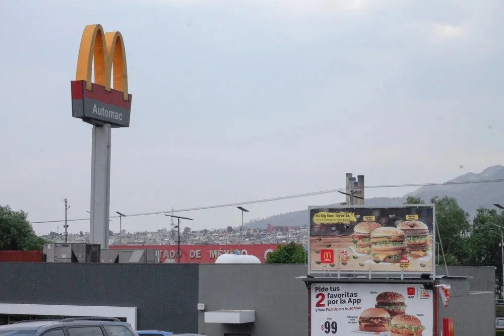 A McDonald's sign with the golden arches and an AutoMac sign below, along with a McDonald's advertisement billboard displaying burgers and promotional information, under a cloudy sky in a cityscape.