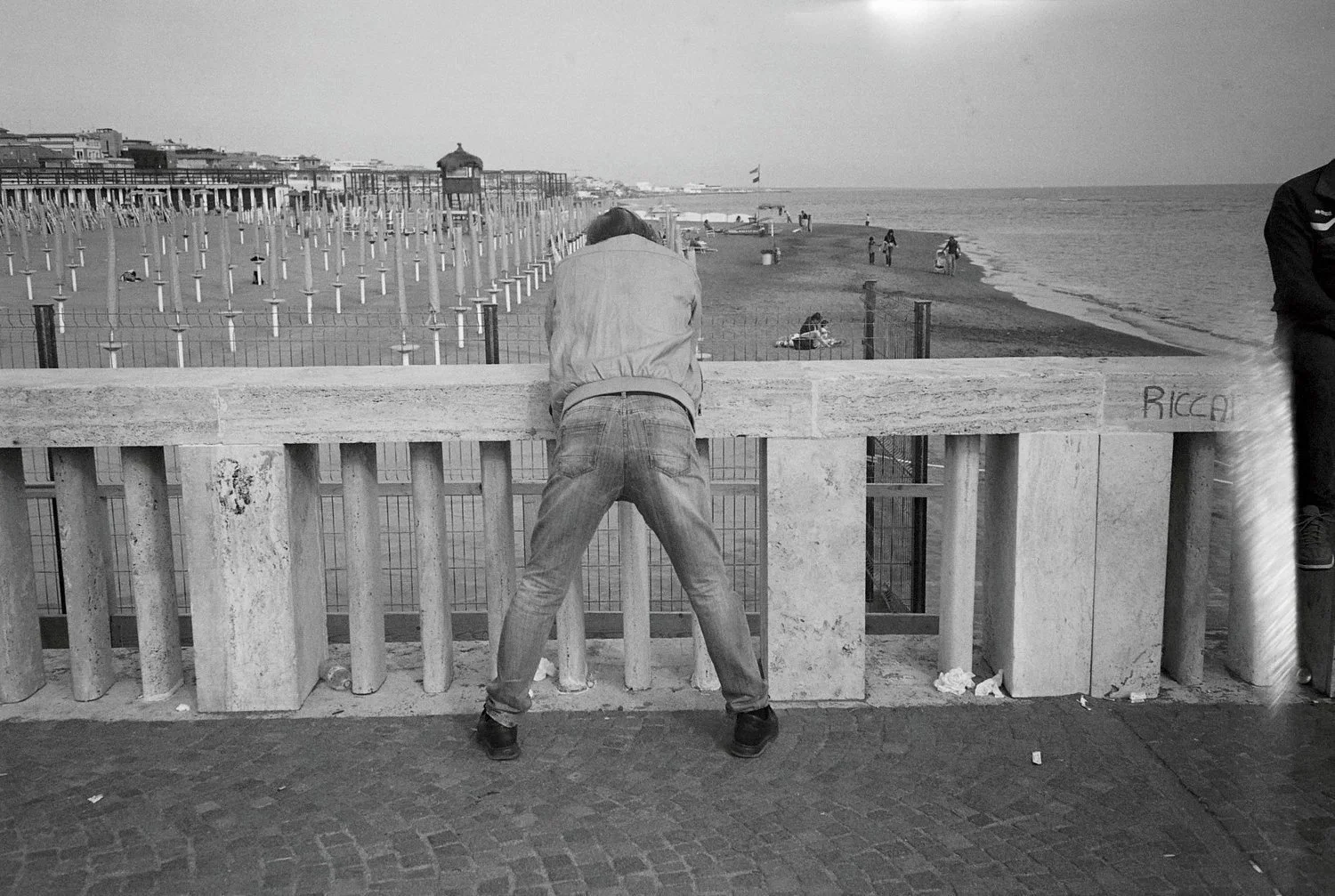 Man leaning on a stone railing, looking out at a beach with umbrellas and a few people, in black and white.