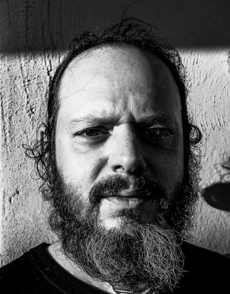 A black and white close-up photo of a man with a beard, long hair, and intense facial expression, standing against a rough wall.