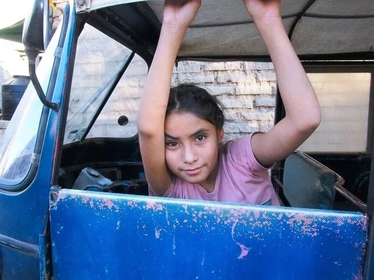A young girl in a pink shirt leaning out of a blue vehicle, holding onto the roof with both hands, looking at the camera with a slight smile.