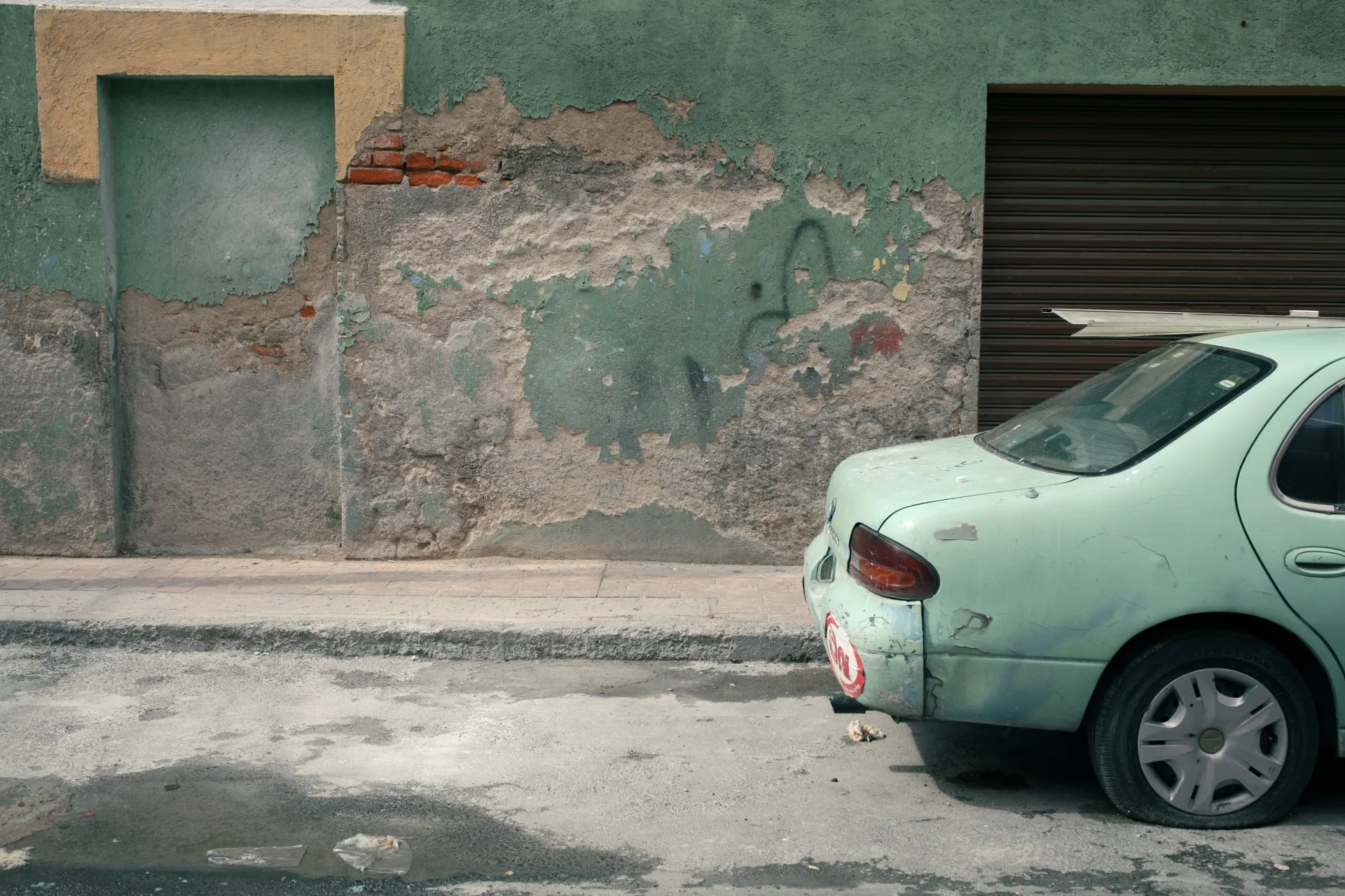 Older mint green car with visible scratches parked on the street in front of a worn wall with peeling paint and graffiti, alongside a closed dark brown garage door.