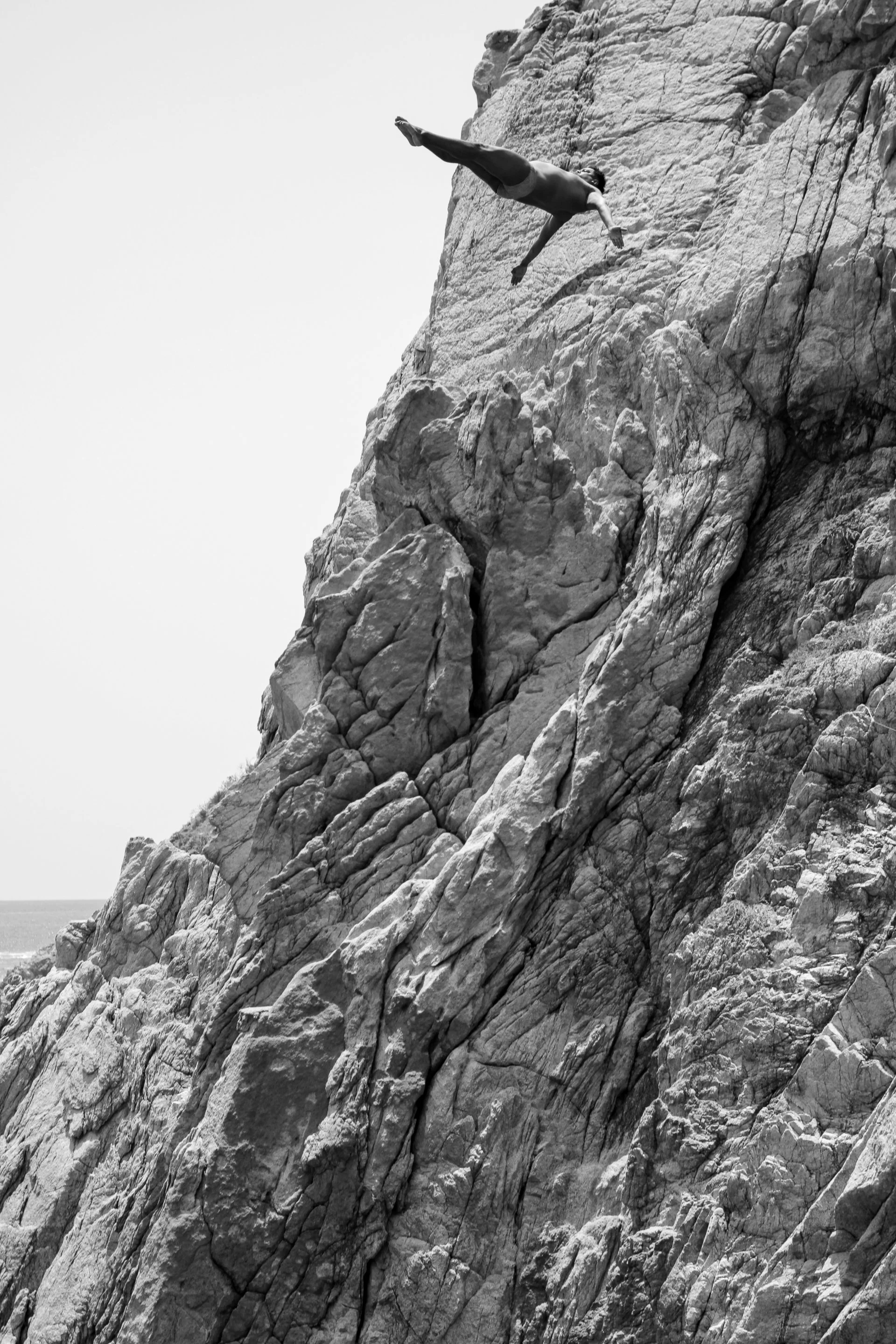 A person rock climbing on a steep, rugged cliffside, gripping the rock with both hands and feet, with clear sky and ocean visible in the background.