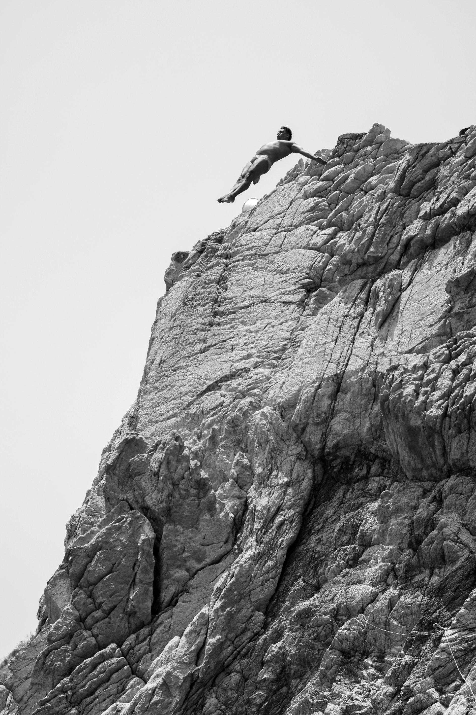A man in swimming trunks is balancing on the edge of a rocky cliff, with his arms outstretched for balance, against a plain sky background.