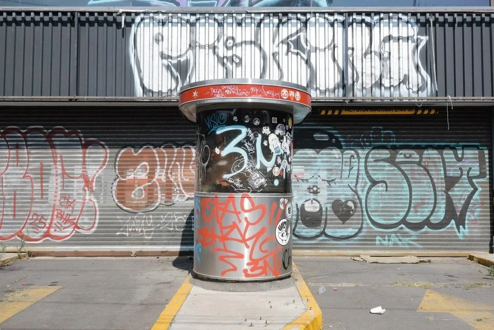 A cylindrical structure covered in graffiti in front of a metal shutter with graffiti, on a concrete lot with yellow parking lines.