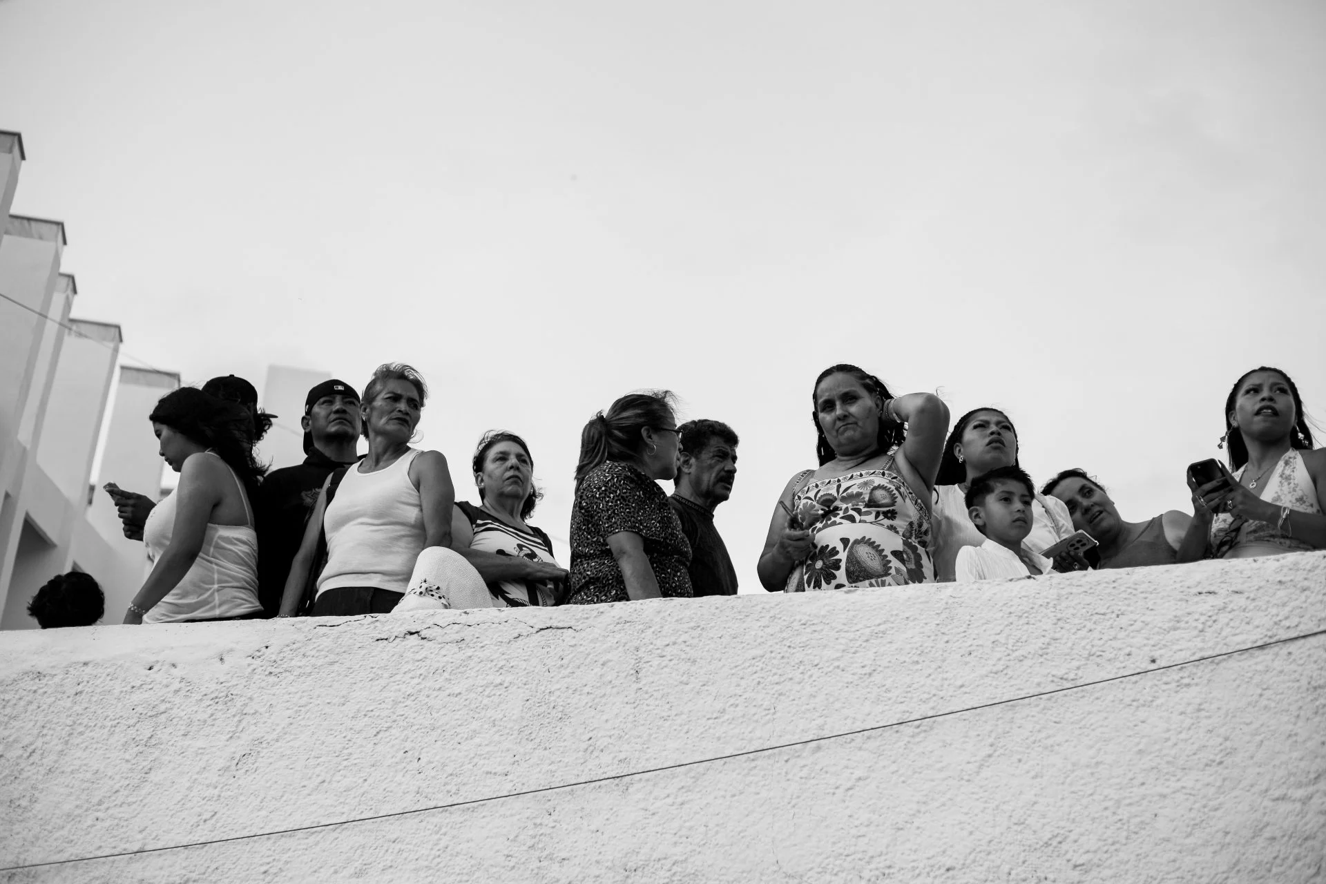 A group of diverse people standing on a balcony, looking in different directions, some talking on phones, and one woman adjusting her hair, with an overcast sky in the background.