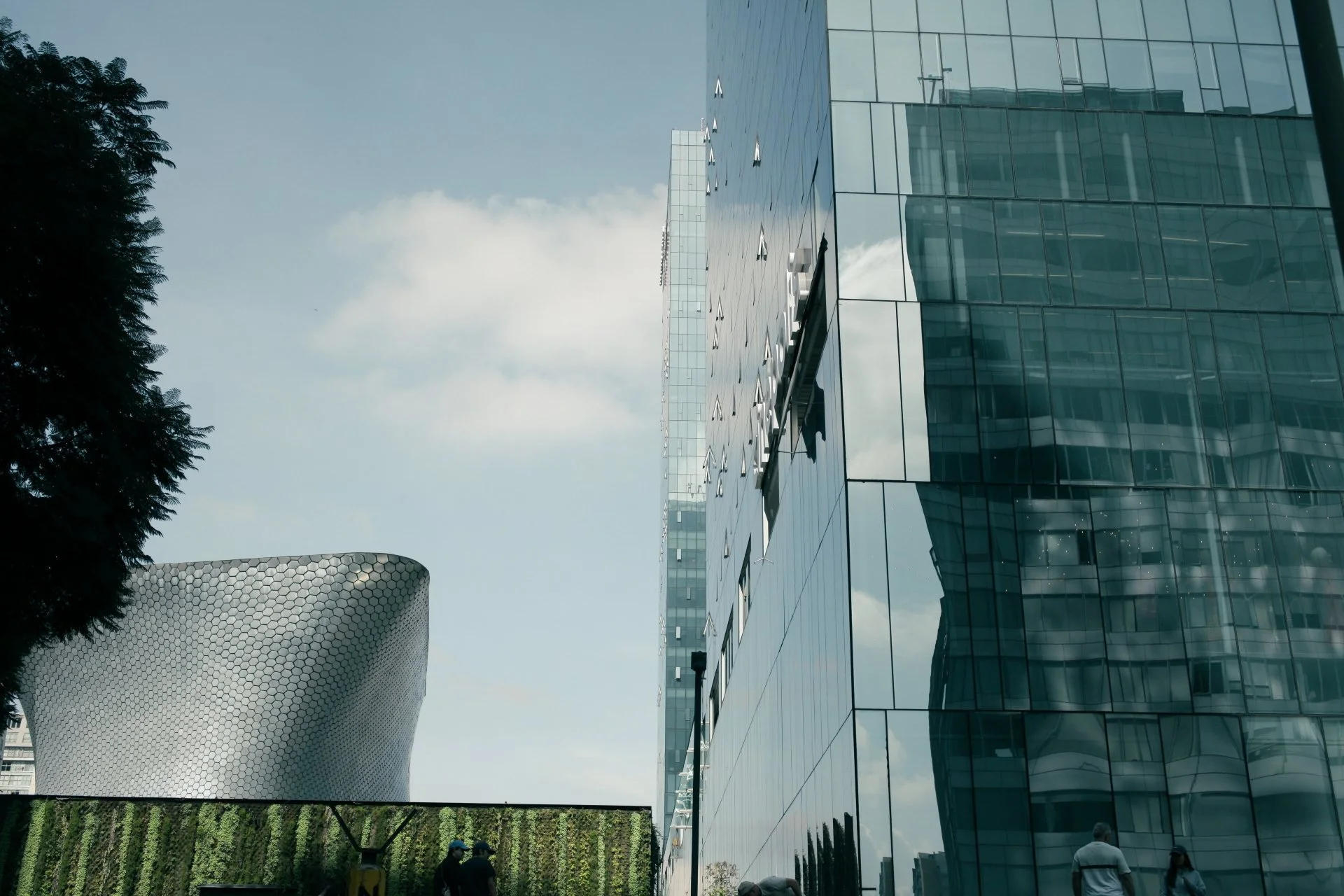 A modern cityscape with a large reflective glass building on the right and a uniquely shaped metallic structure on the left. There are a few people visible at the bottom of the image near a vertical garden wall.