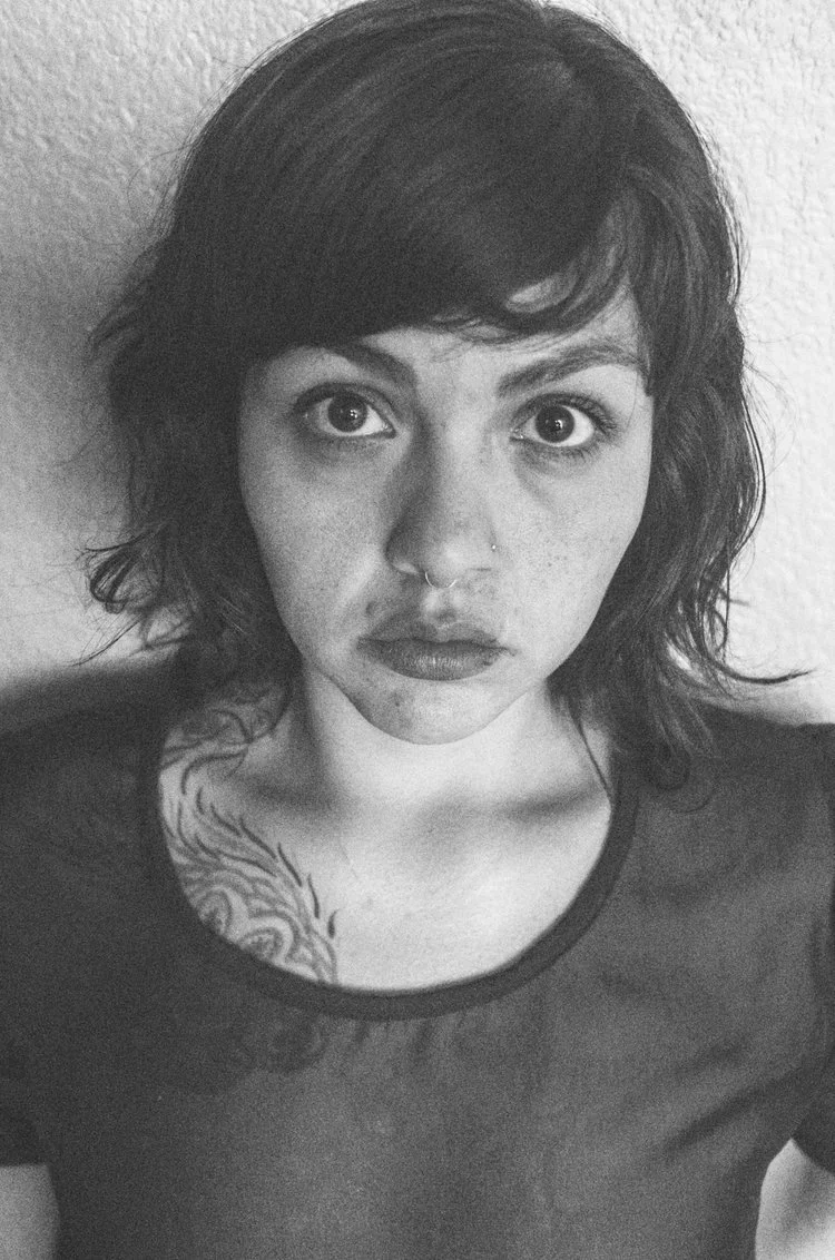 Black and white portrait of a woman with short wavy hair, a septum piercing, and a tattoo on her shoulder, looking directly at the camera.